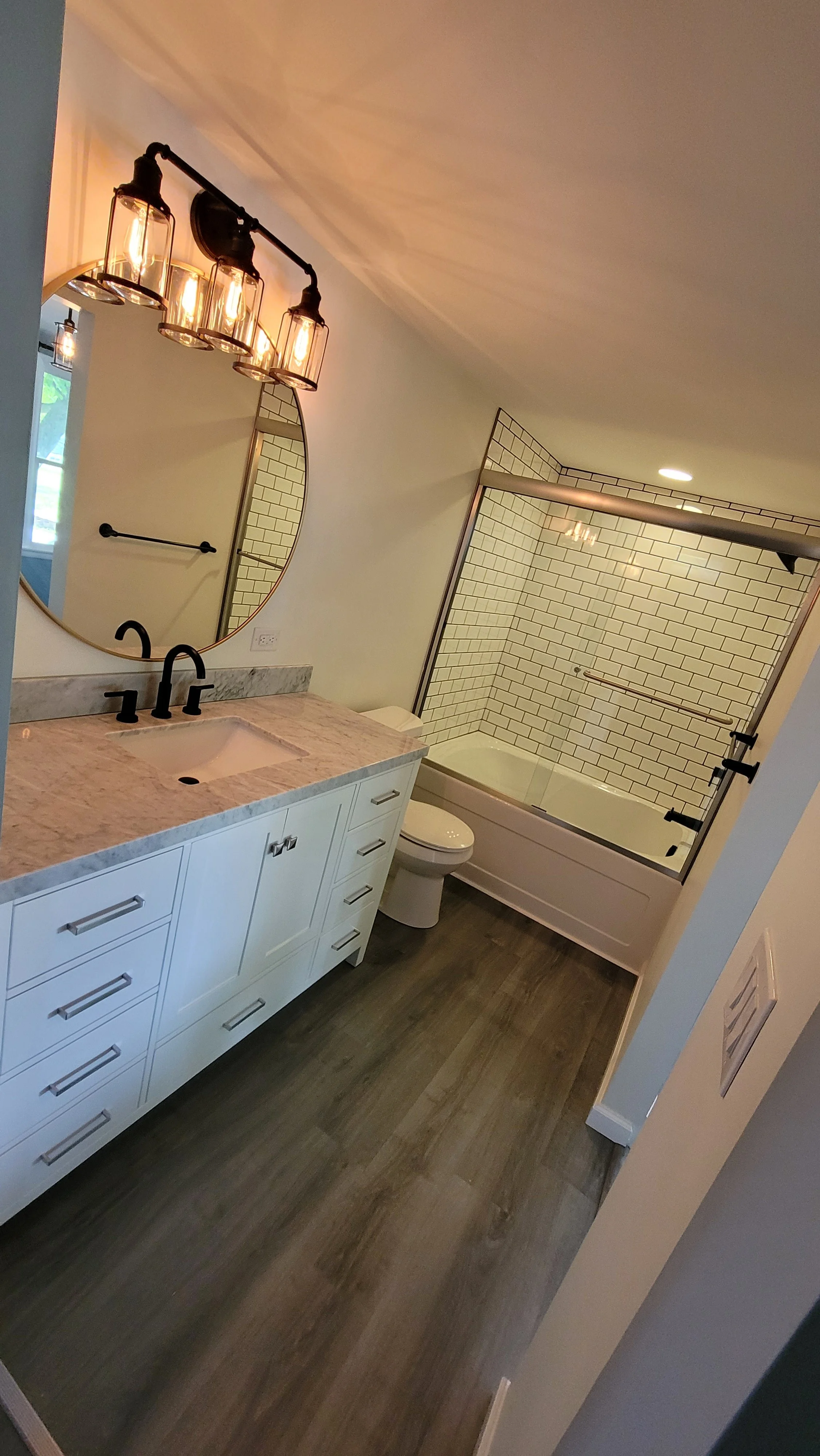 A modern bathroom with a white vanity, marble countertop, black faucet, round mirror, black light fixture, in-wall toilet, and a bathtub with white subway tile walls and a glass door.