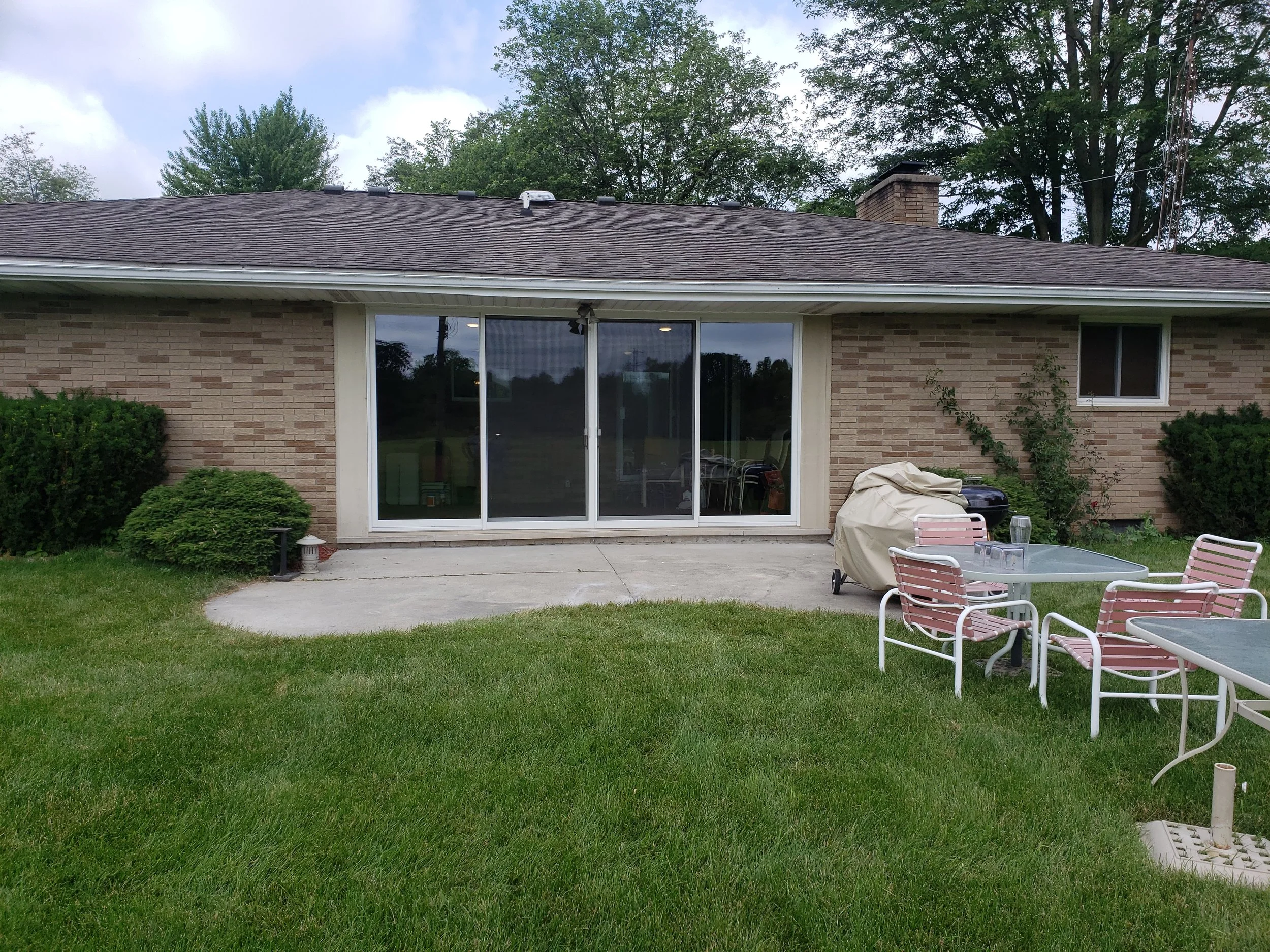 Backyard patio with lawn chairs, covered grill, sliding glass door, and plants in front of a brick house.