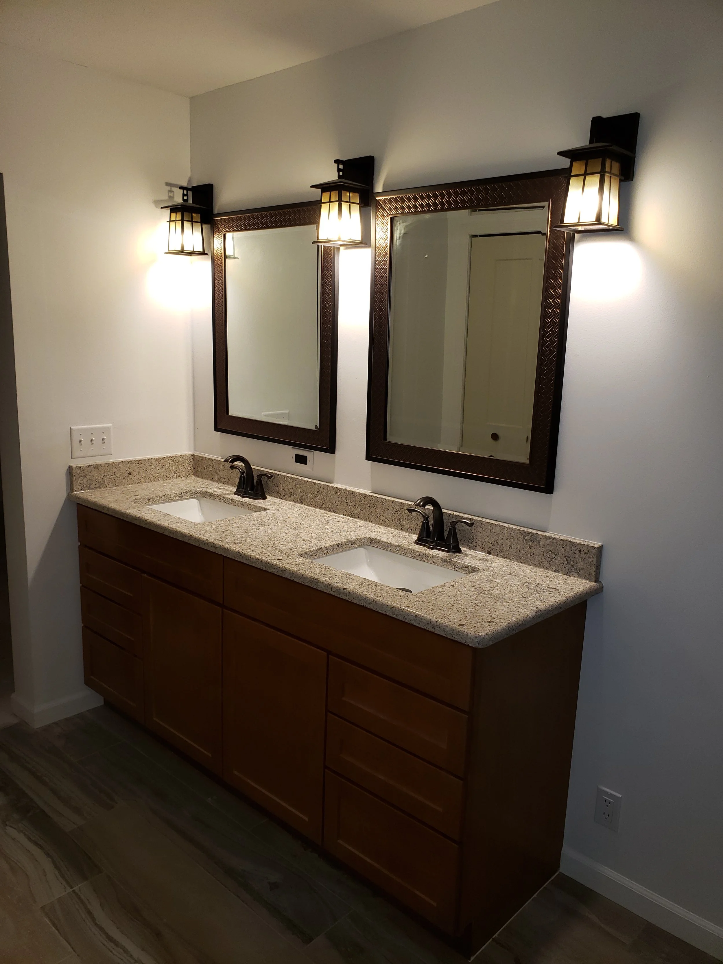 A bathroom vanity area with two mirrors, two wall-mounted light fixtures, a granite countertop with two sinks, a wooden cabinet base, and a tiled floor.