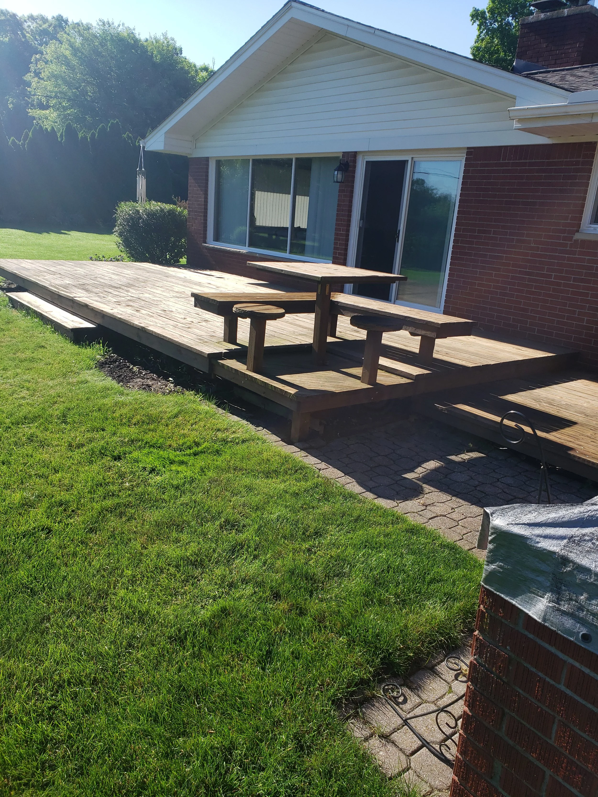 Backyard patio with wooden deck, picnic table, and a brick house with sliding glass door and large window, surrounded by grass and trees.