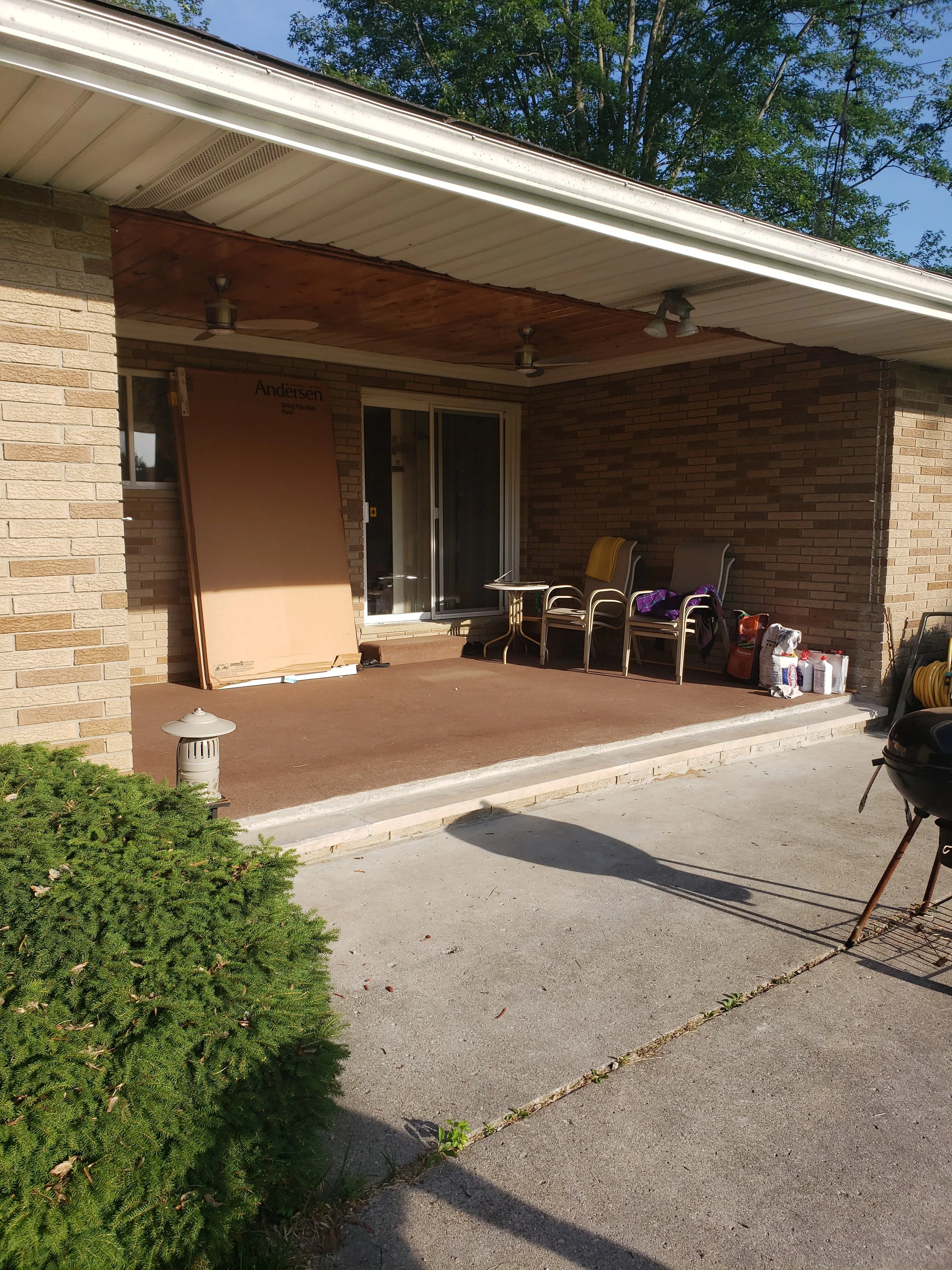 A covered patio area with chairs, a small table, and gardening supplies. The house has brick walls and a wooden ceiling with ceiling fans, and there is greenery and a concrete path in the foreground.