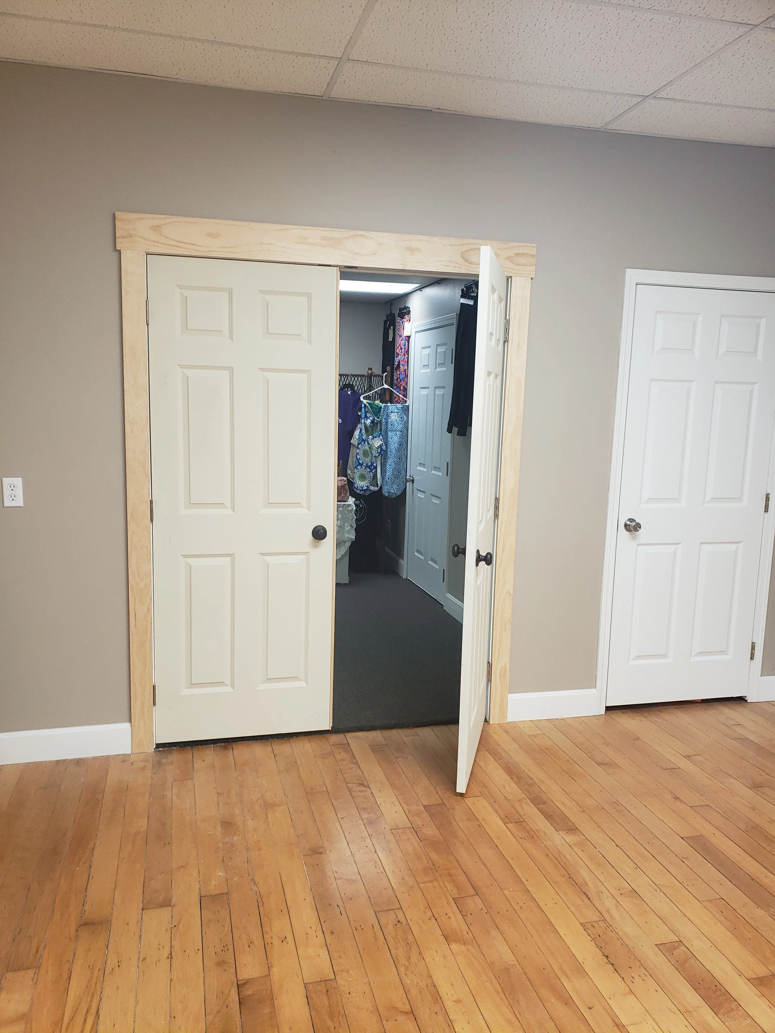 A room with wooden flooring, beige painted walls, and white doors. One door is partly open, revealing a closet with hanging clothes and various items. The ceiling has acoustic tiles.