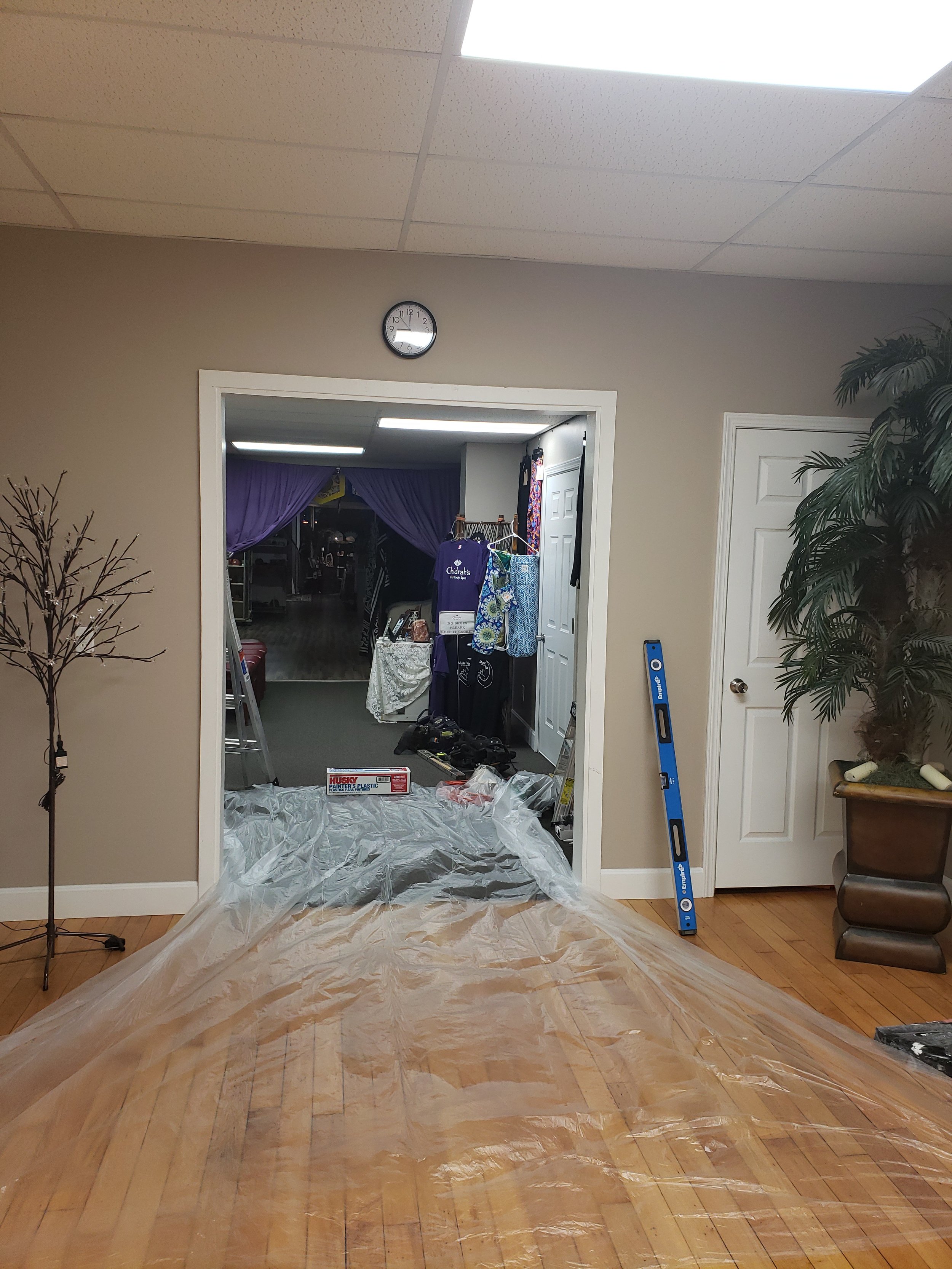 Inside a room undergoing renovation, with a plastic drop cloth on the wooden floor, a partially visible aluminum ladder, and a doorway leading to another room with purple curtains, various tools, and materials stored inside.