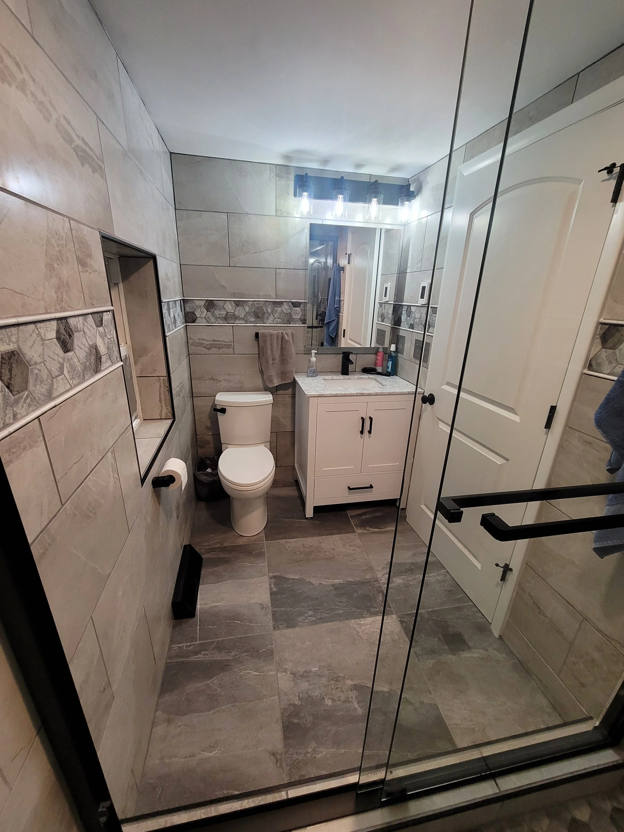 A modern bathroom with gray and beige tile on the walls and floor, a white vanity with black hardware, a mirror, a toilet, and towels. There is a door and a glass shower door reflected in the mirror.