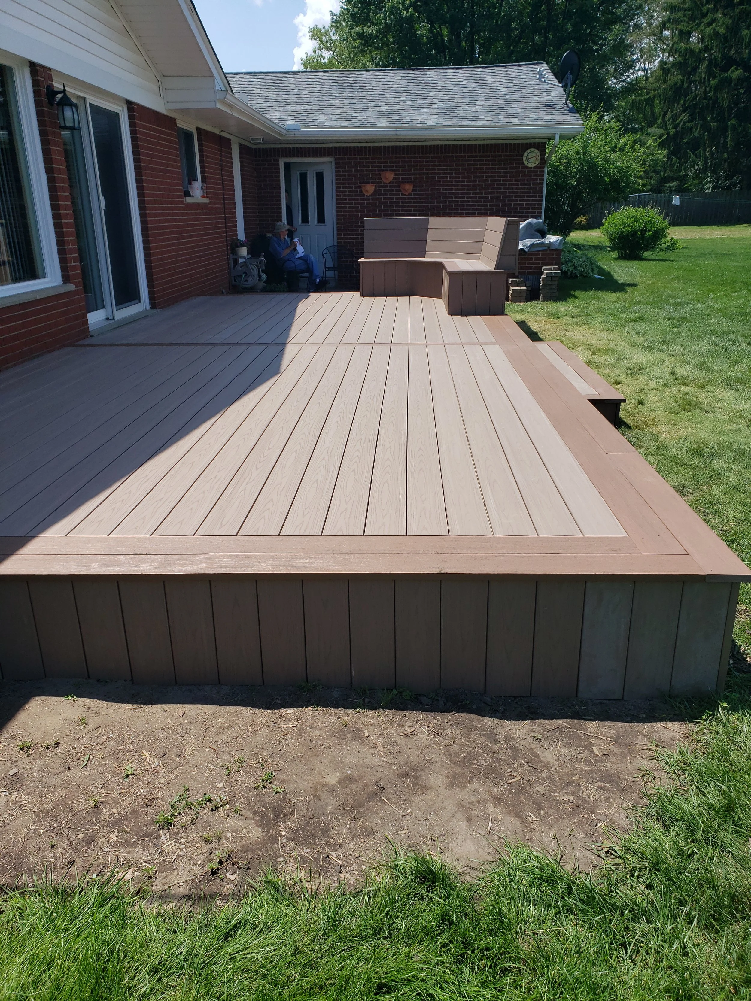 A backyard with a newly installed wooden deck attached to a brick house. The deck has a built-in bench and is bordered by grass and dirt. There is a woman sitting on a chair near the house, looking at her phone. In the background, there are trees, bushes, and a wooden fence.