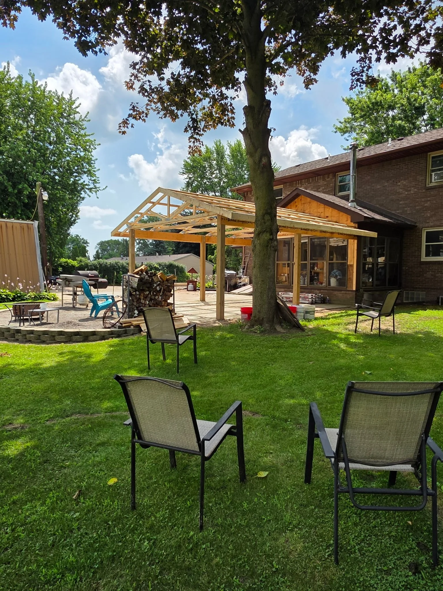 Backyard with chairs, a large tree, a partially constructed wooden patio cover, and a brick house under a partly cloudy sky.