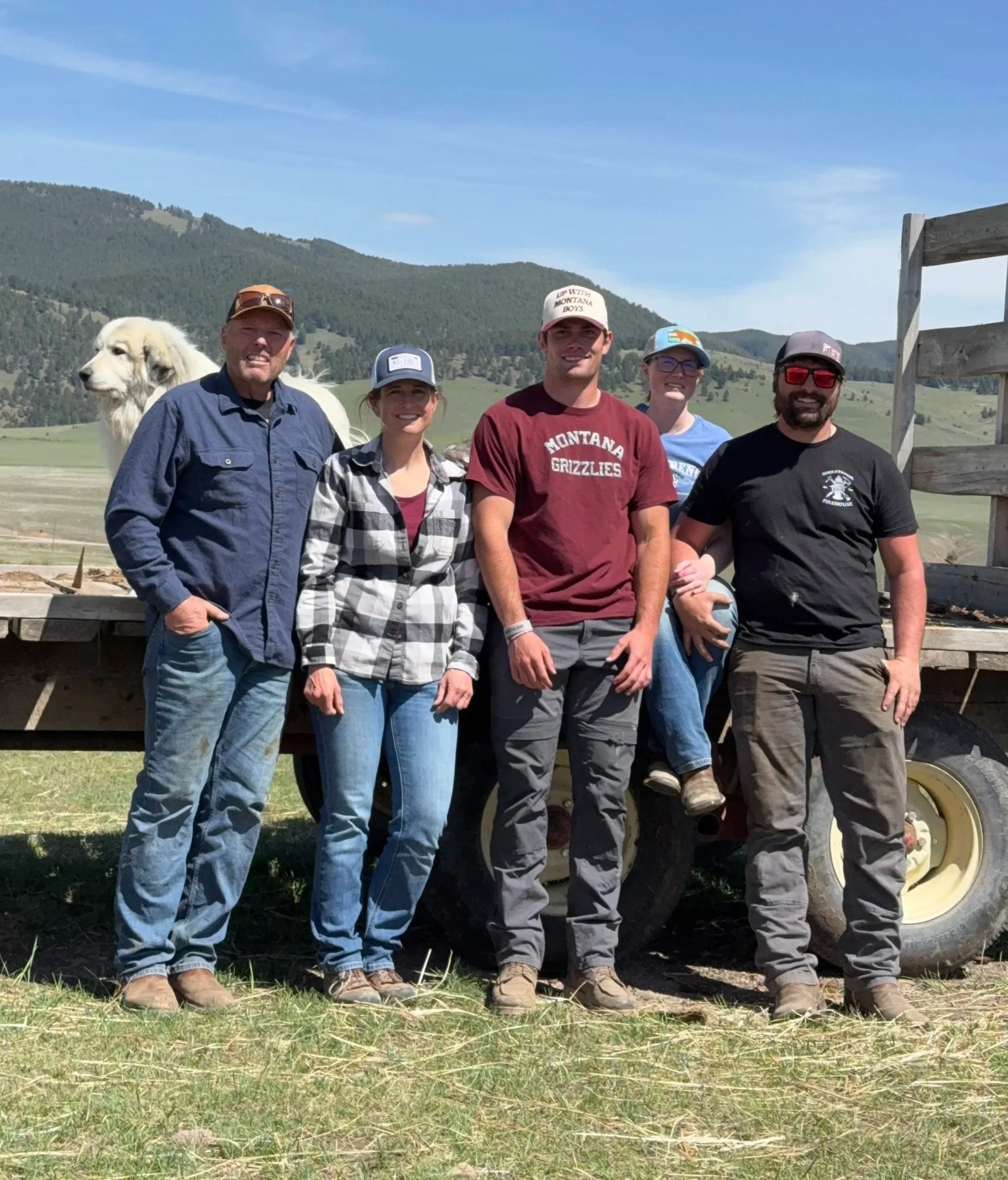 A group of five people standing in front of a tractor on a farm with green hills and mountains in the background under a blue sky. A white dog is visible behind the group.