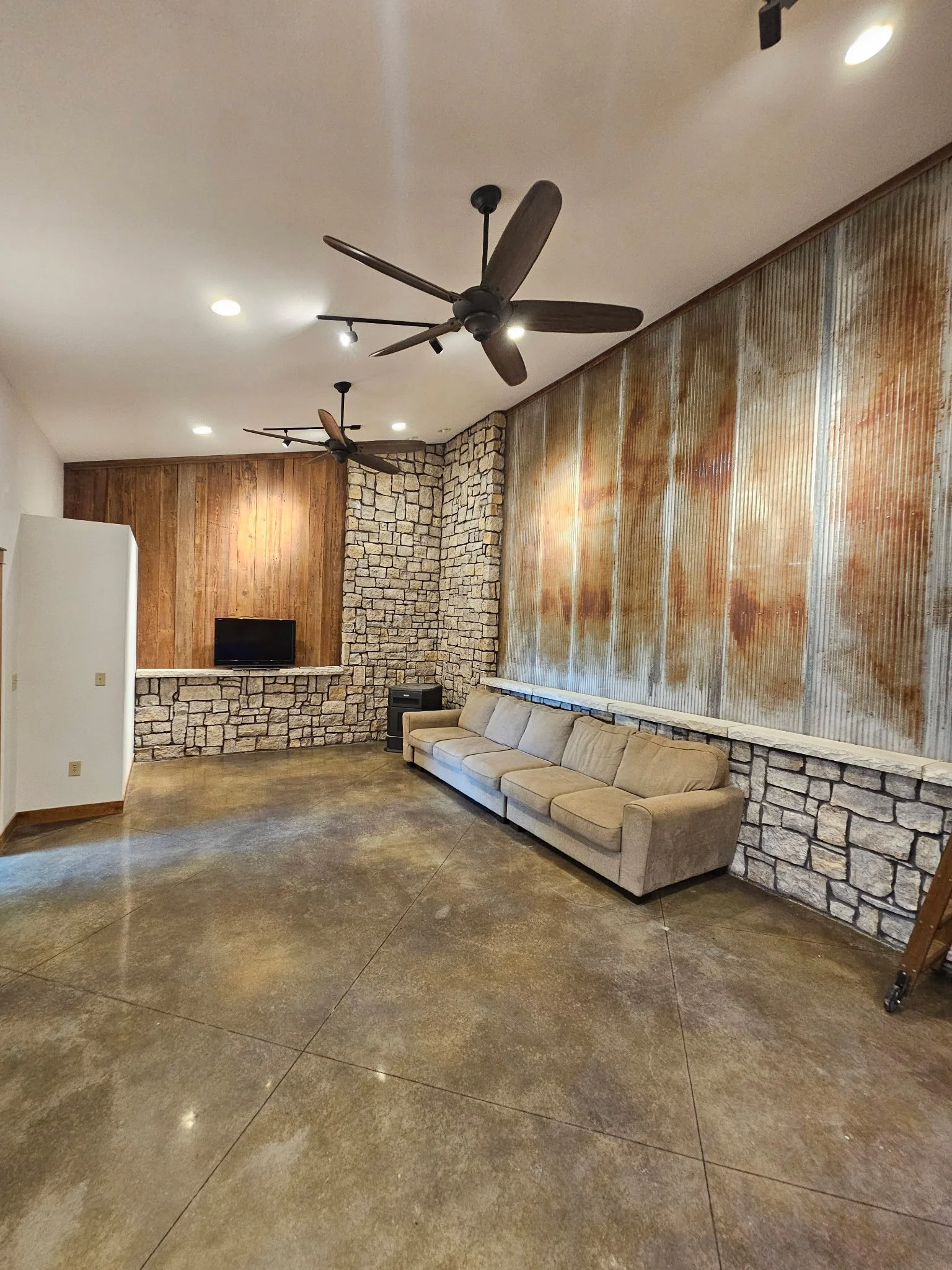 Interior of a room with a stone and wood wall, a beige sectional sofa, a small TV on a stone ledge, and ceiling fans with wooden blades.