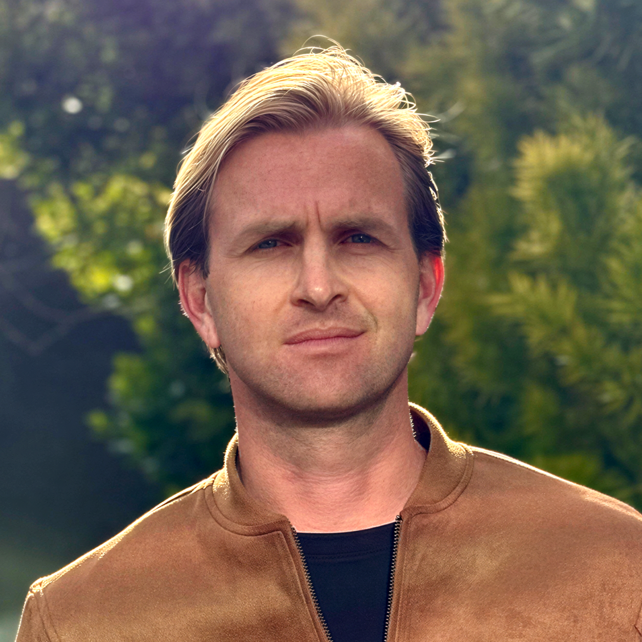 Portrait of a man outdoors with trees and greenery in the background, wearing a brown jacket and black shirt, with sunlight highlighting his hair.