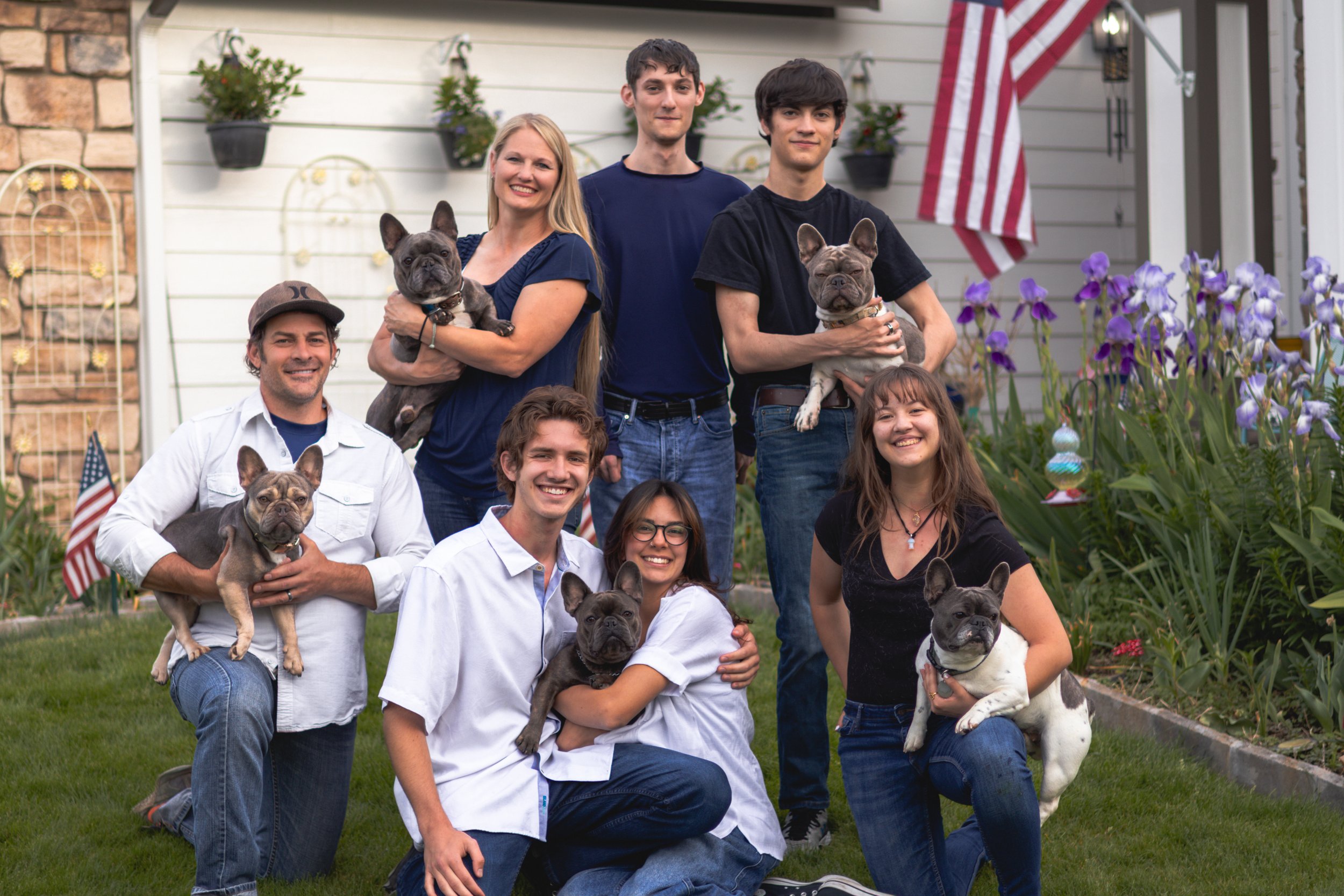 A group of ten people, including children and adults, along with five French Bulldogs, posing in a garden in front of a house with American flags, flowers, and potted plants.