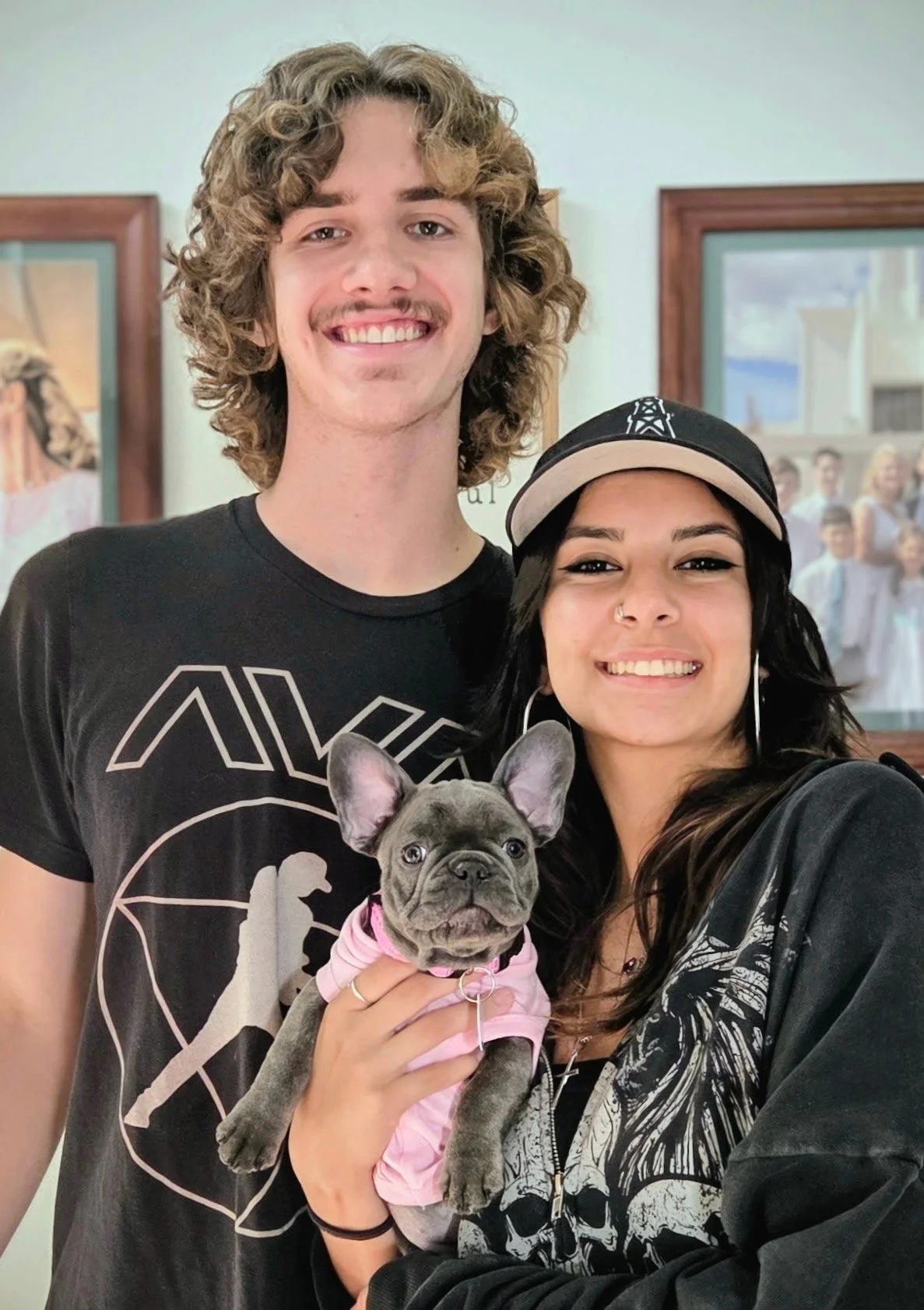 A young man with curly hair and a girl with dark hair wearing a baseball cap, smiling and holding a French Bulldog puppy in a pink outfit, indoors with framed pictures in the background.