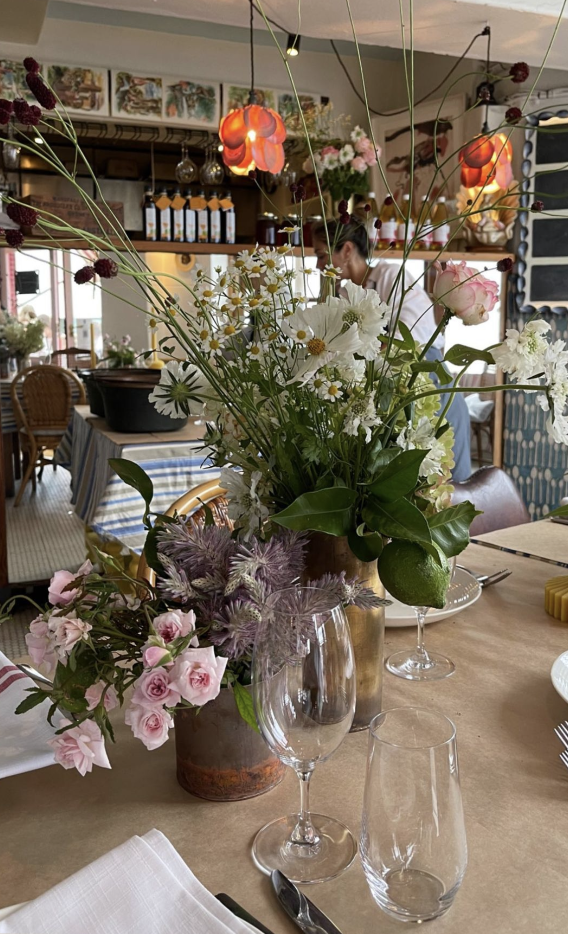 A close-up view of a table set with floral centerpieces, wine glasses, and cutlery in a cozy restaurant or cafe with warm lighting and decorative artwork on the walls.