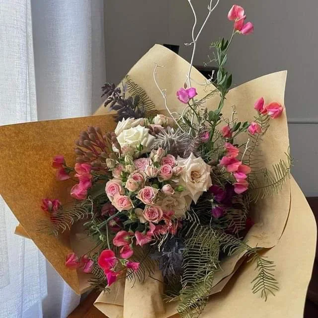 A bouquet of pink and white roses, pink sweet peas, fern leaves, and other greenery wrapped in tan and beige paper.