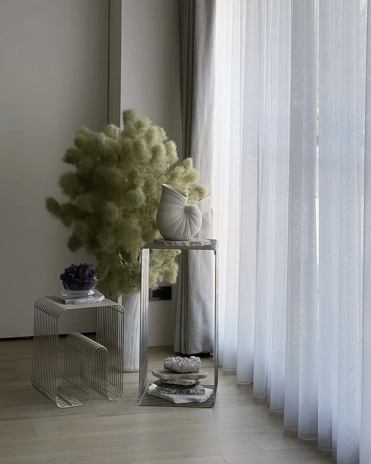 Interior with sheer curtains, a decorative shell-shaped vase on a metal stand, a large beige pampas grass arrangement, a purple flower in a glass bowl, and crystal cluster on a small metallic table.