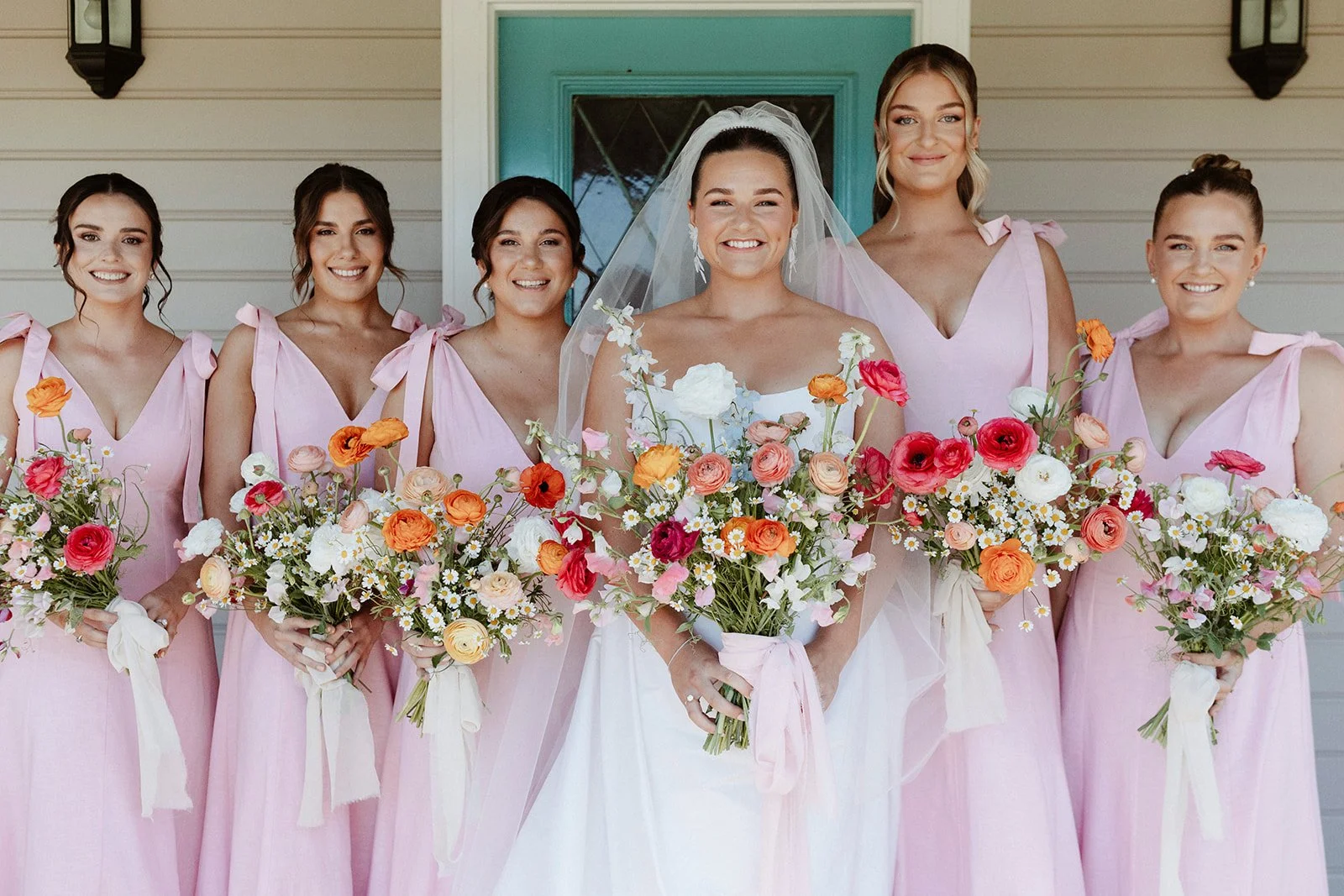 Bride and bridesmaids standing in front of a house, holding colorful flower bouquets. The bride is in the center wearing a wedding dress and veil.