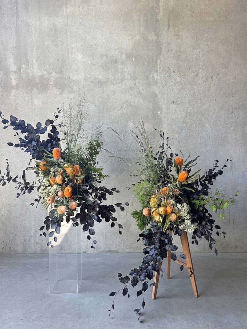 Two floral arrangements with dark purple leaves, white flowers, and peach-colored berries, one on a clear acrylic stand and the other on a wooden easel, against a plain gray wall.
