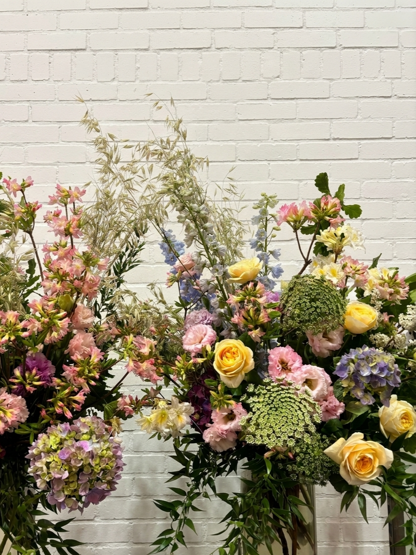 A colorful floral arrangement with pink, yellow, purple, and white flowers set against a white brick wall.