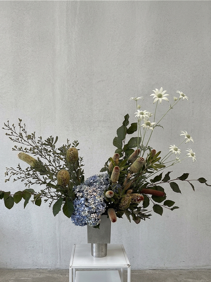 A large floral arrangement in a metallic vase on a white stand against a plain light gray wall. The arrangement includes white daisies, blue hydrangeas, and various green foliage and textured plants.