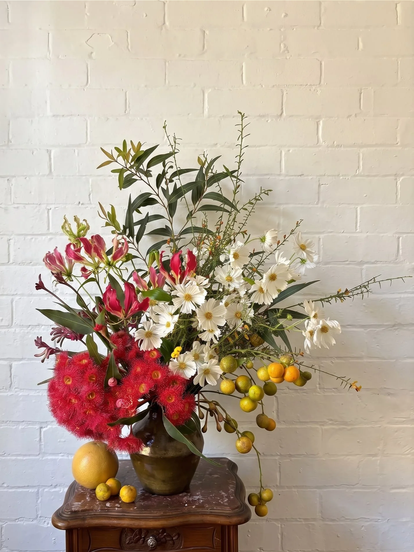 A floral arrangement in a dark brown vase on a wooden table, against a white brick wall. The bouquet includes pink flowers, white daisies, red fluffy flowers, and yellowish-green berries, with some lemons placed on the table.