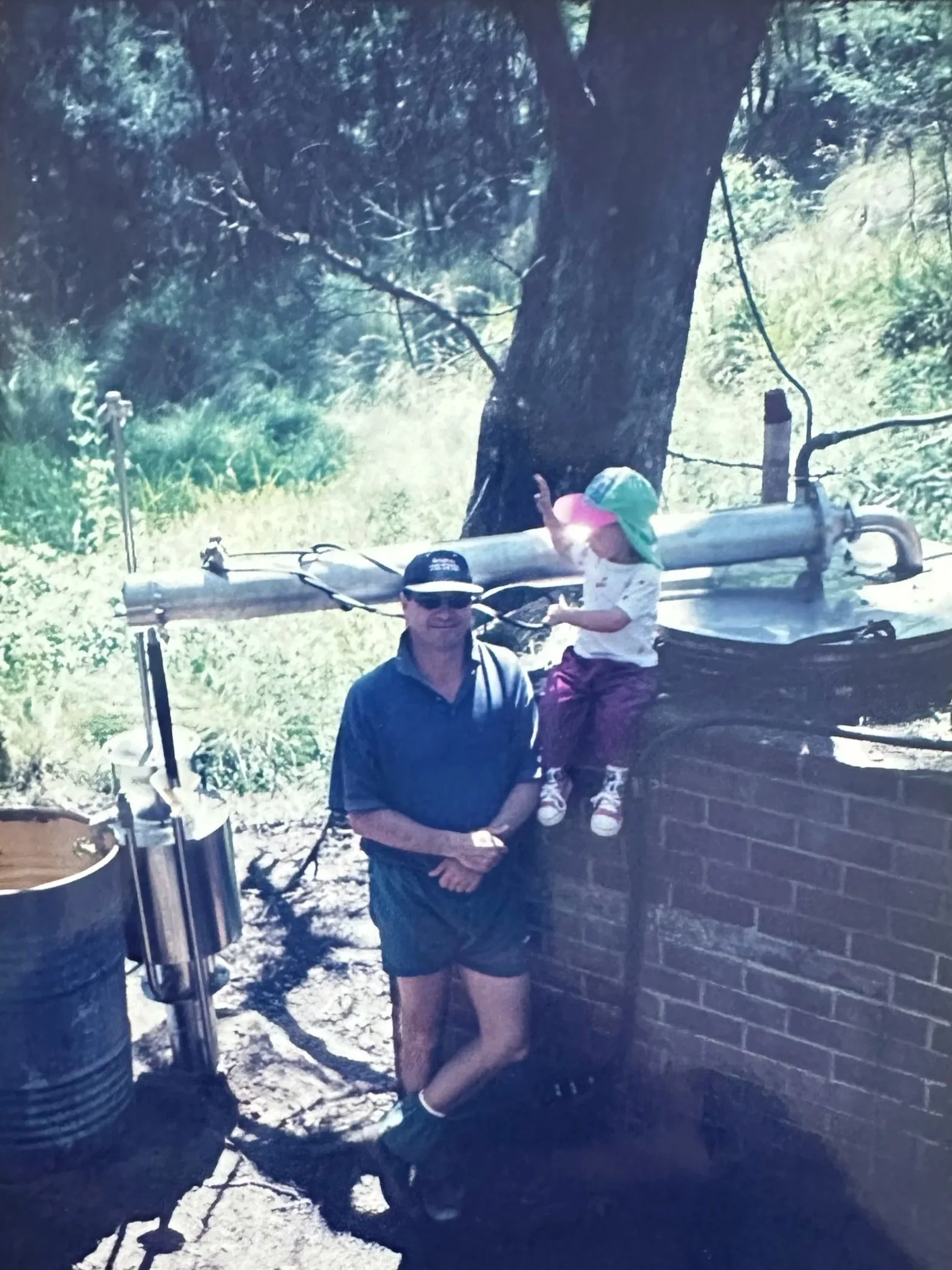 A man and a young girl, possibly a family, enjoying outdoor time near a large tree and a brick wall. The man is wearing a dark shirt and a cap, standing next to a water tank or barrel. The girl, wearing a sun hat, is sitting on the brick wall, with a large metal pipe or utility structure behind her. The scene appears to be in a sunny, rural or semi-rural area with some greenery and a gravel or dirt ground.