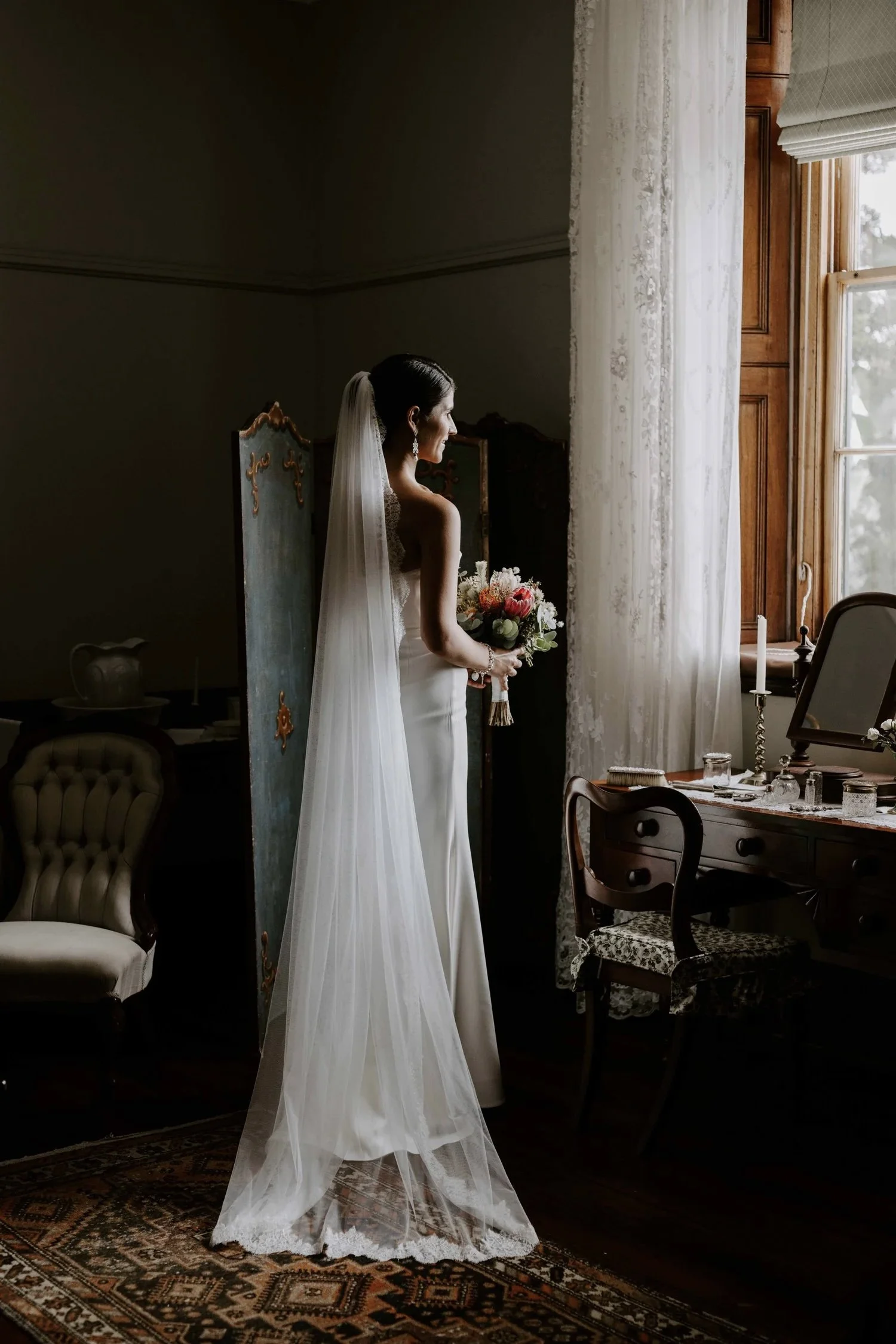 Bride standing indoors near a large window, wearing a white wedding dress and veil, holding a bouquet, in a vintage-style room with antique furniture and a patterned rug.