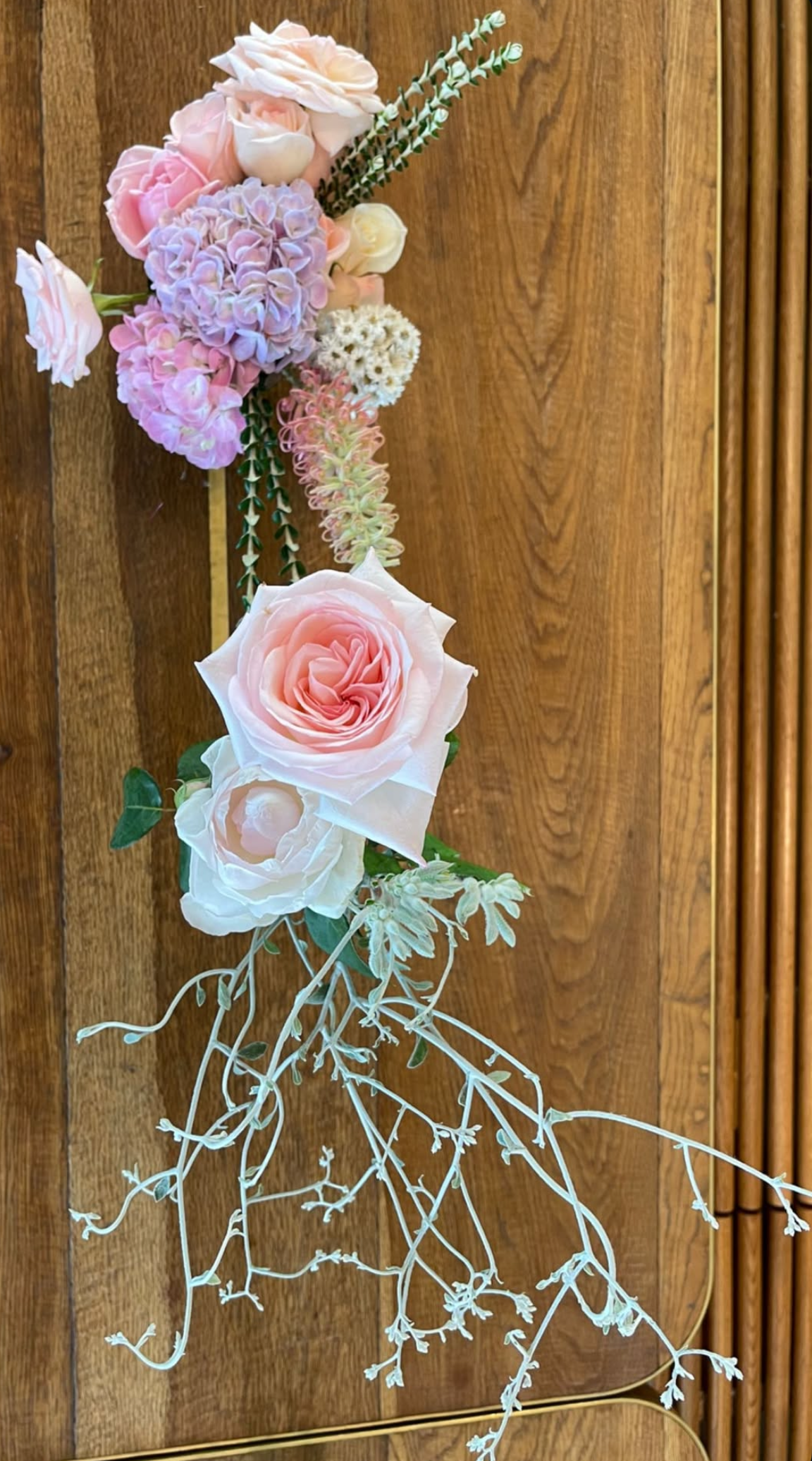 Artificial floral arrangement featuring pink roses, purple hydrangeas, white flowers, and green foliage on a wooden background.