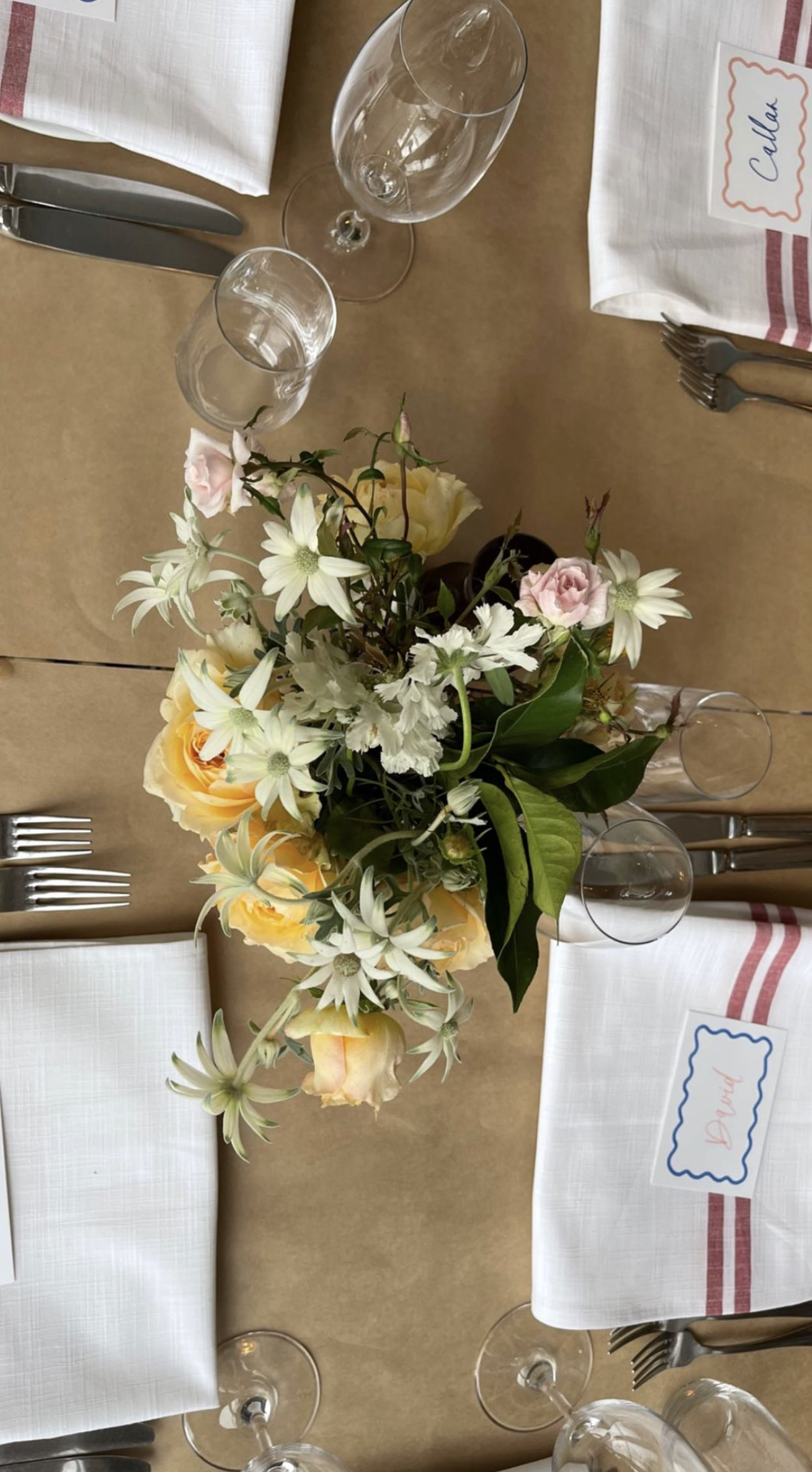A table setting with white cloth napkins, clear wine glasses, forks, and knives, featuring a floral centerpiece with white, yellow, and pink flowers on a brown table.