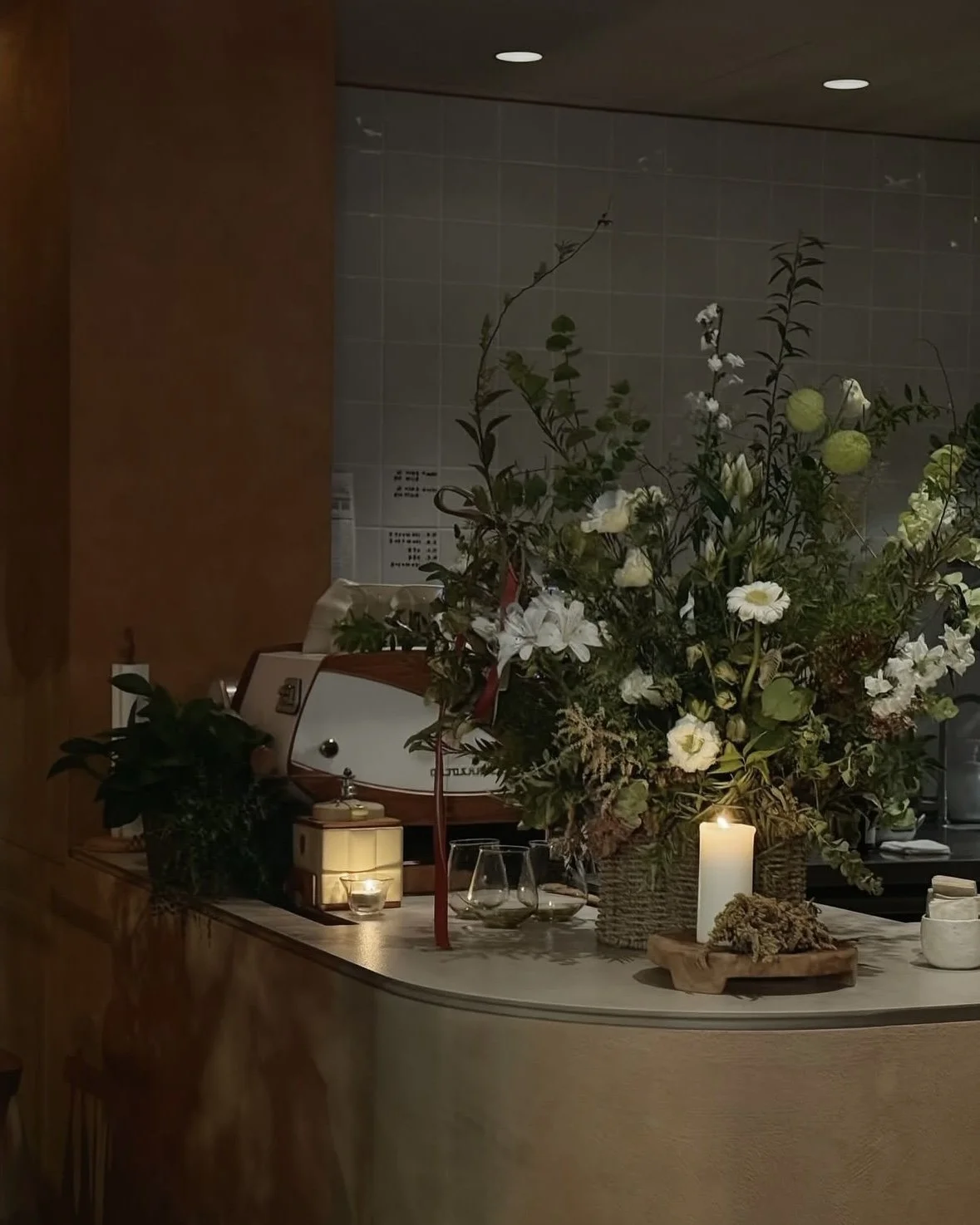 A floral arrangement with white flowers and greenery on a table, lit by candles, in a warm, indoor setting.