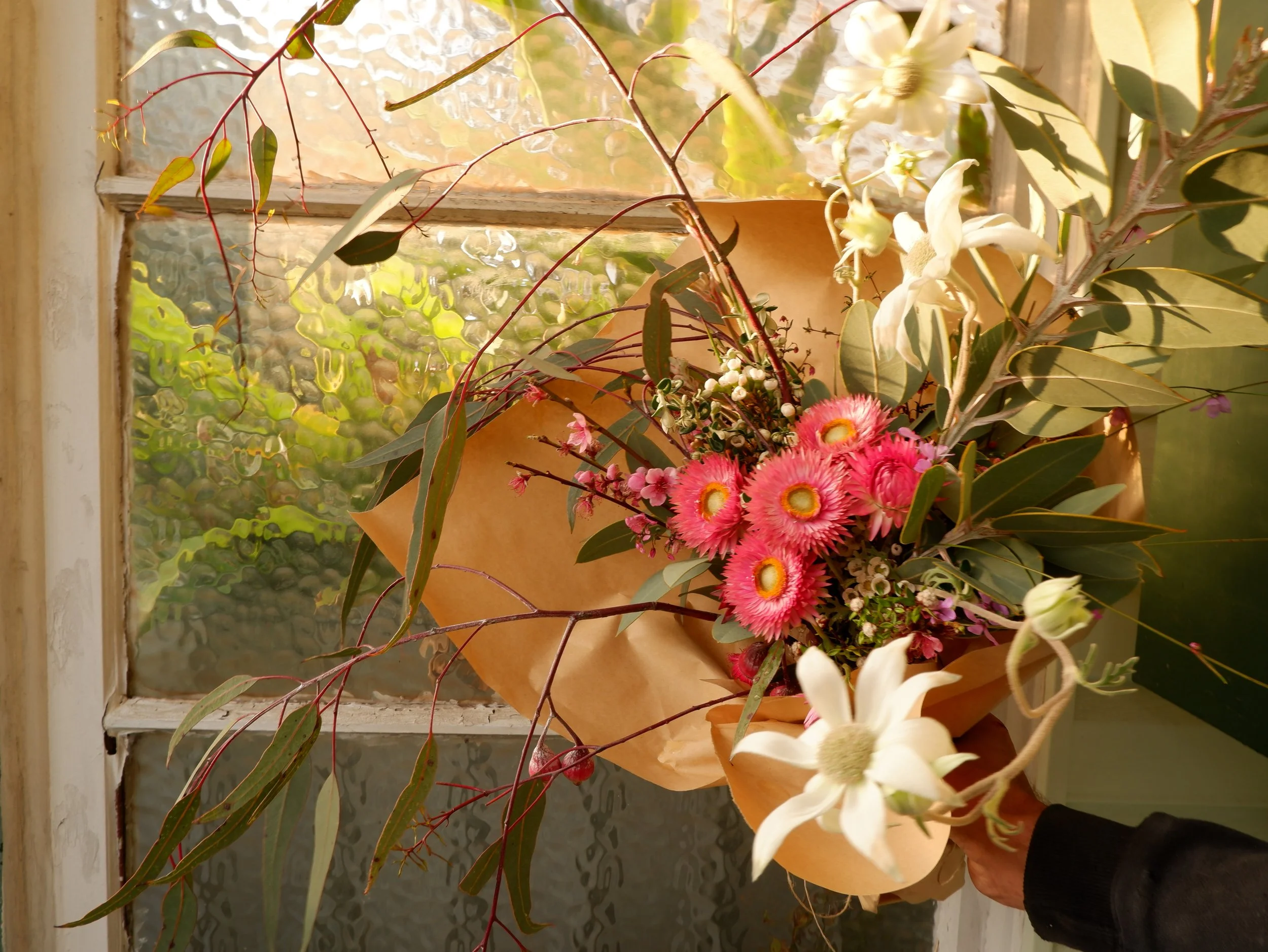 A bouquet of flowers and greenery wrapped in brown paper held by a person's hand in front of a textured window.
