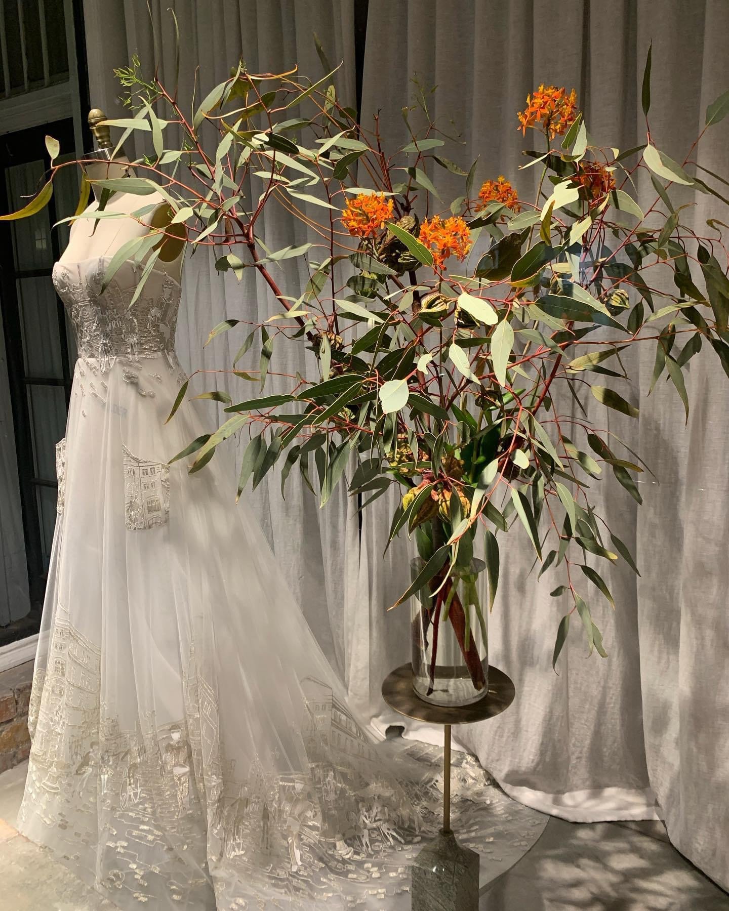 A mannequin wearing a wedding dress with lace details standing behind a large potted plant with orange flowers. The scene is indoors with gray curtains in the background.