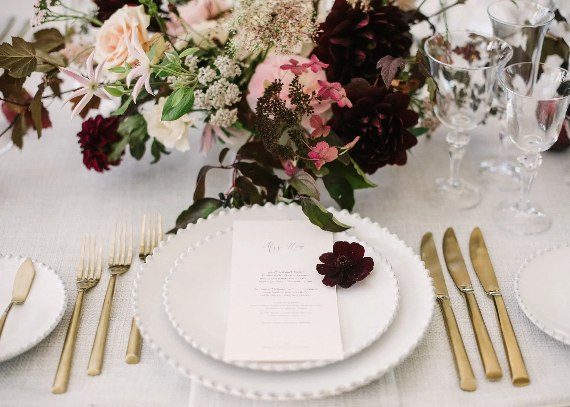 A close-up of a wedding or event tablescape featuring a white plate with a printed menu and a dark red flower, gold cutlery, and a floral centerpiece with pink, white, and dark red flowers and greenery, along with empty clear glassware.