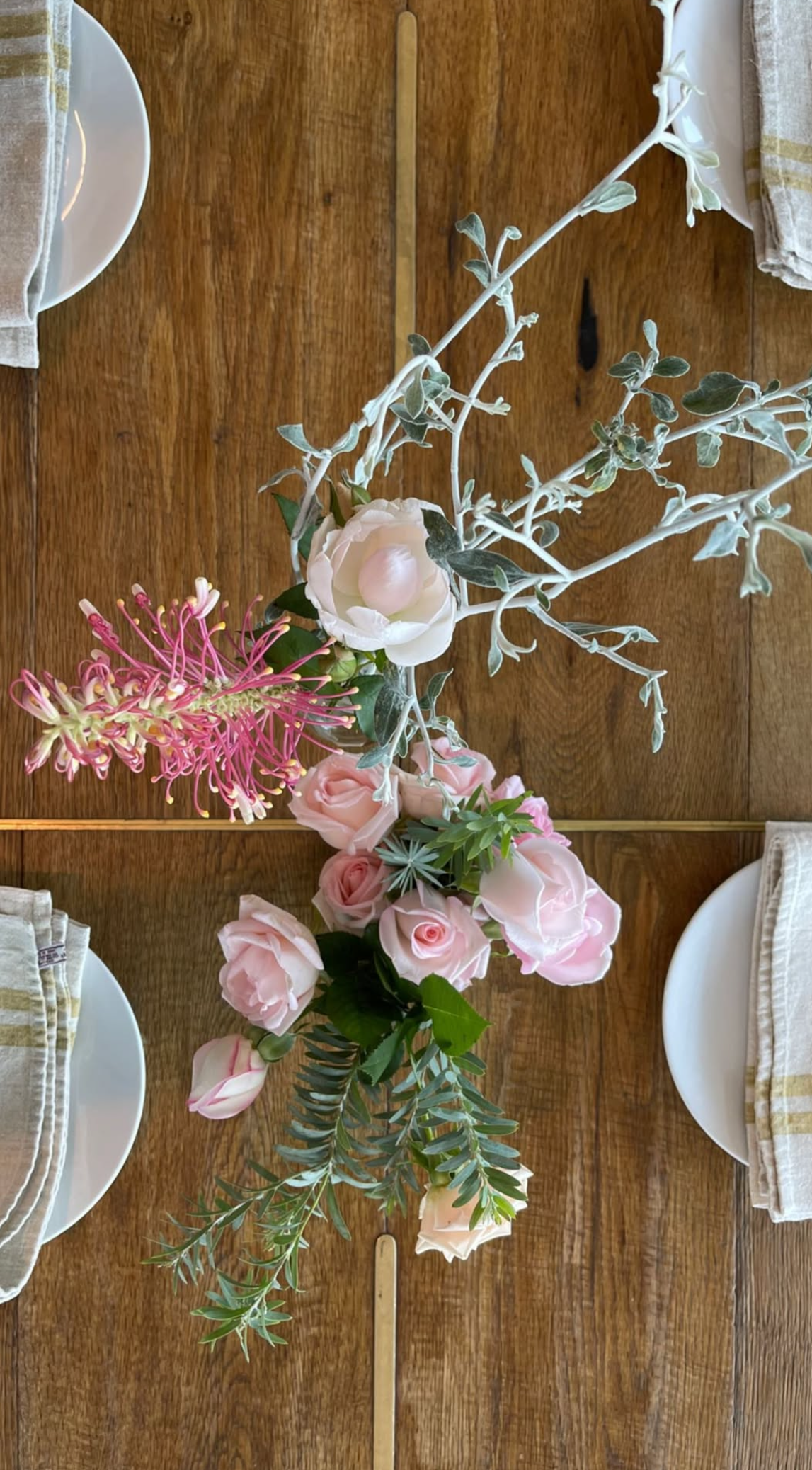 A floral centerpiece on a wooden dining table with pink roses, white roses, greenery, and a pink flower with long, thin petals.