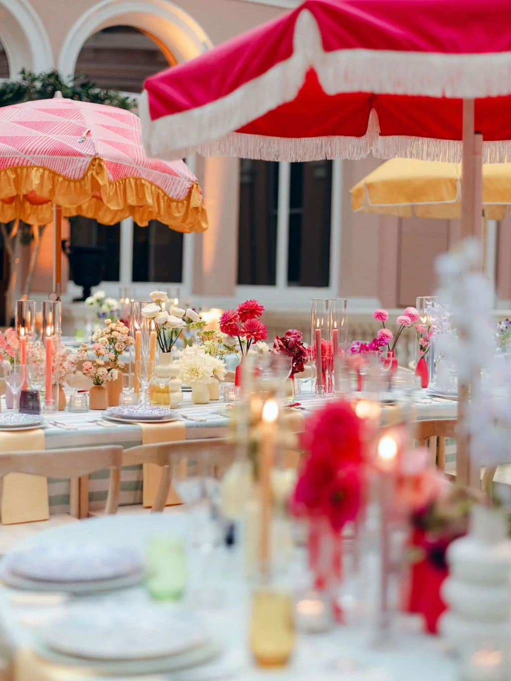 Outdoor dining setup with tables, colorful umbrellas, and floral arrangements with pink, white, and red flowers, candles, and tableware.