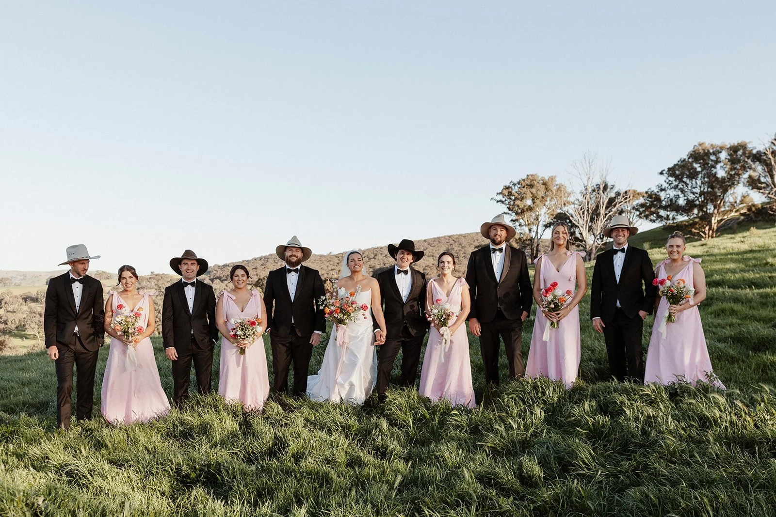 A wedding party standing outdoors on green grass with hills and trees in the background, dressed in formal attire, some women holding bouquets, in the late afternoon sunlight.
