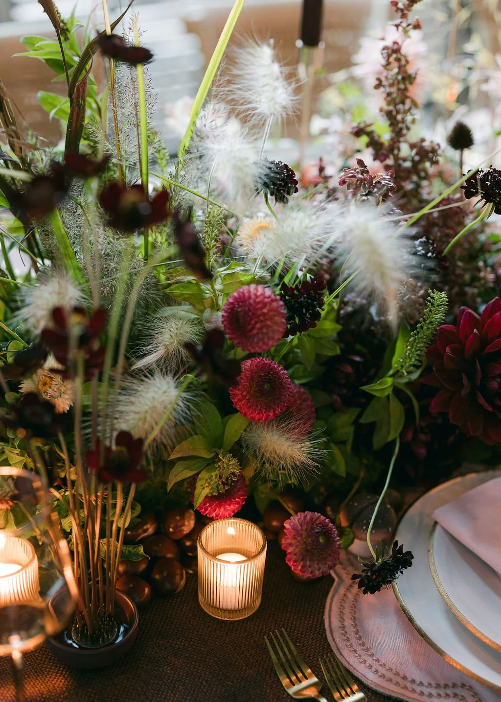 A table setting with a large floral arrangement of various colorful flowers, including purple, red, and white blooms, complemented by small candle holders with lit candles, and a neatly arranged white plate and napkin on the right side.