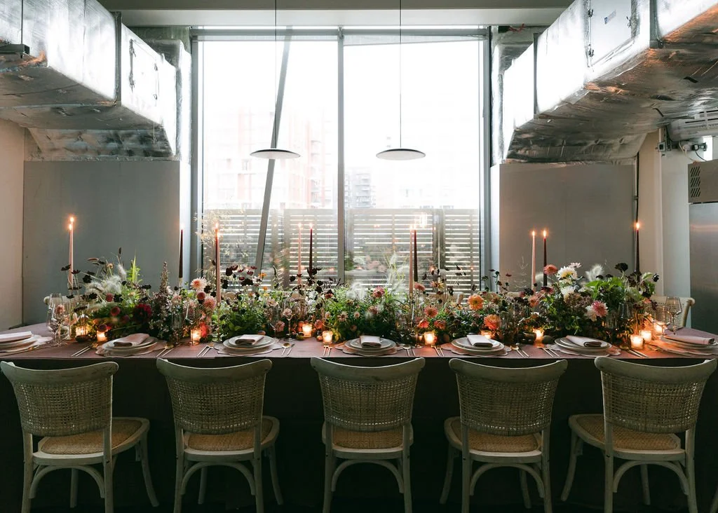 A long dining table decorated with flowers, candles, and place settings, set up for a special event or dinner in a modern, urban space with large windows and natural light.