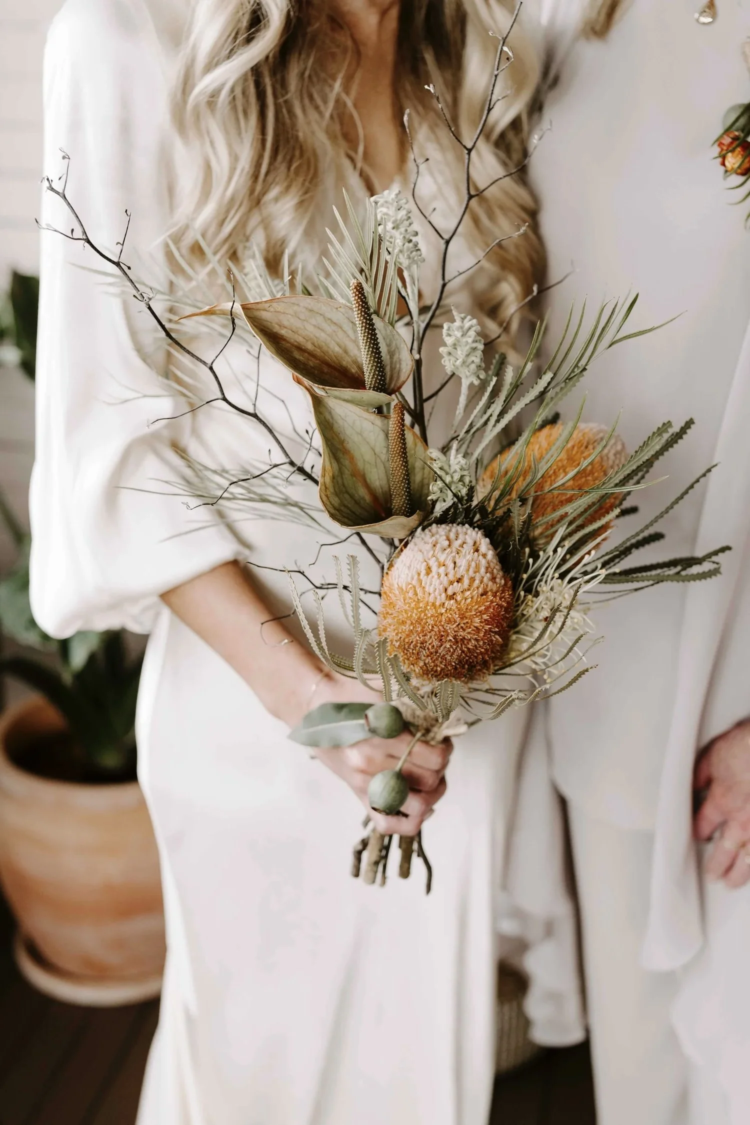 Person holding a bouquet of dried flowers and foliage, including palm leaves, branches, and textured blooms, wearing a white outfit.