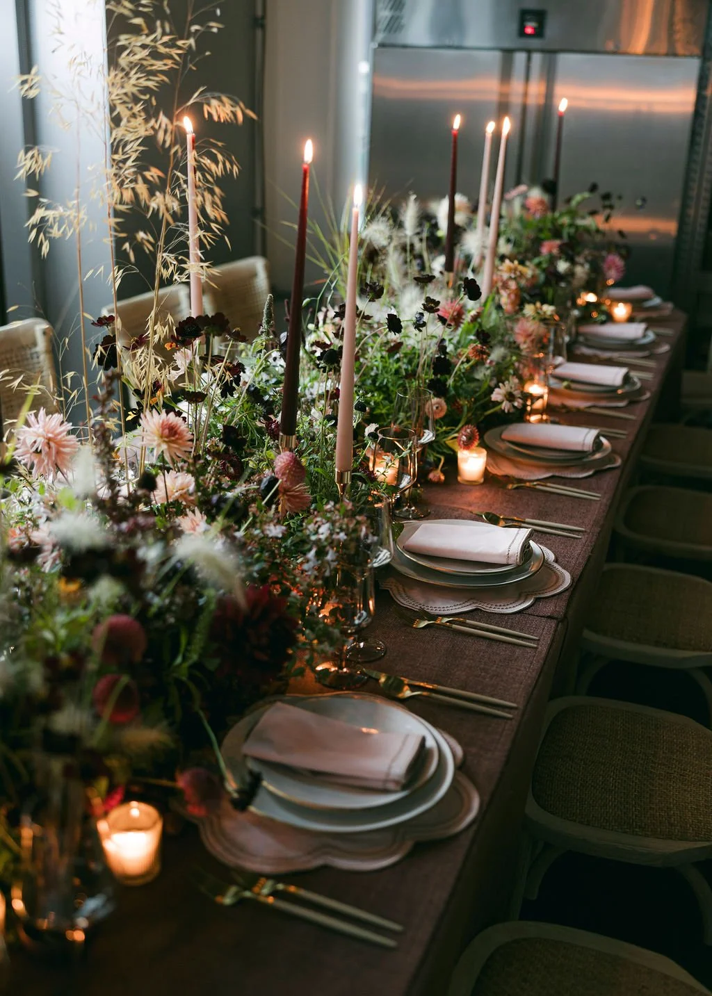 A long dining table decorated with a lush floral centerpiece, tall pink and white candles, and small votive candles, set with white plates, gold utensils, and white napkins for a formal event.