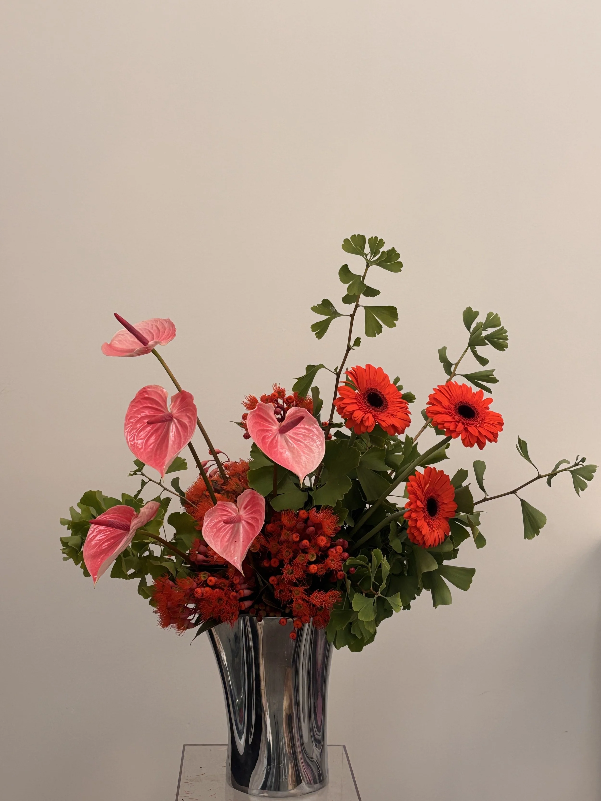 A floral arrangement in a tall, reflective silver vase with pink anthuriums, orange gerbera daisies, and green foliage, set against a plain off-white background.
