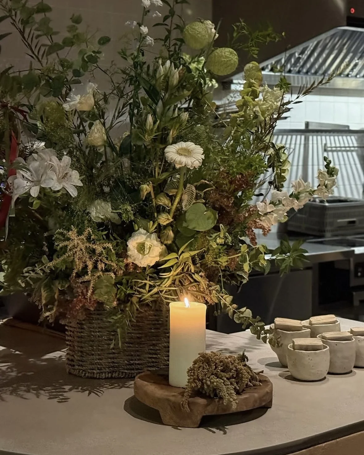 A floral arrangement in a wicker basket, a lit white candle on a wooden tray, and small ceramic cups on a table with a neutral cloth, set against a kitchen background.