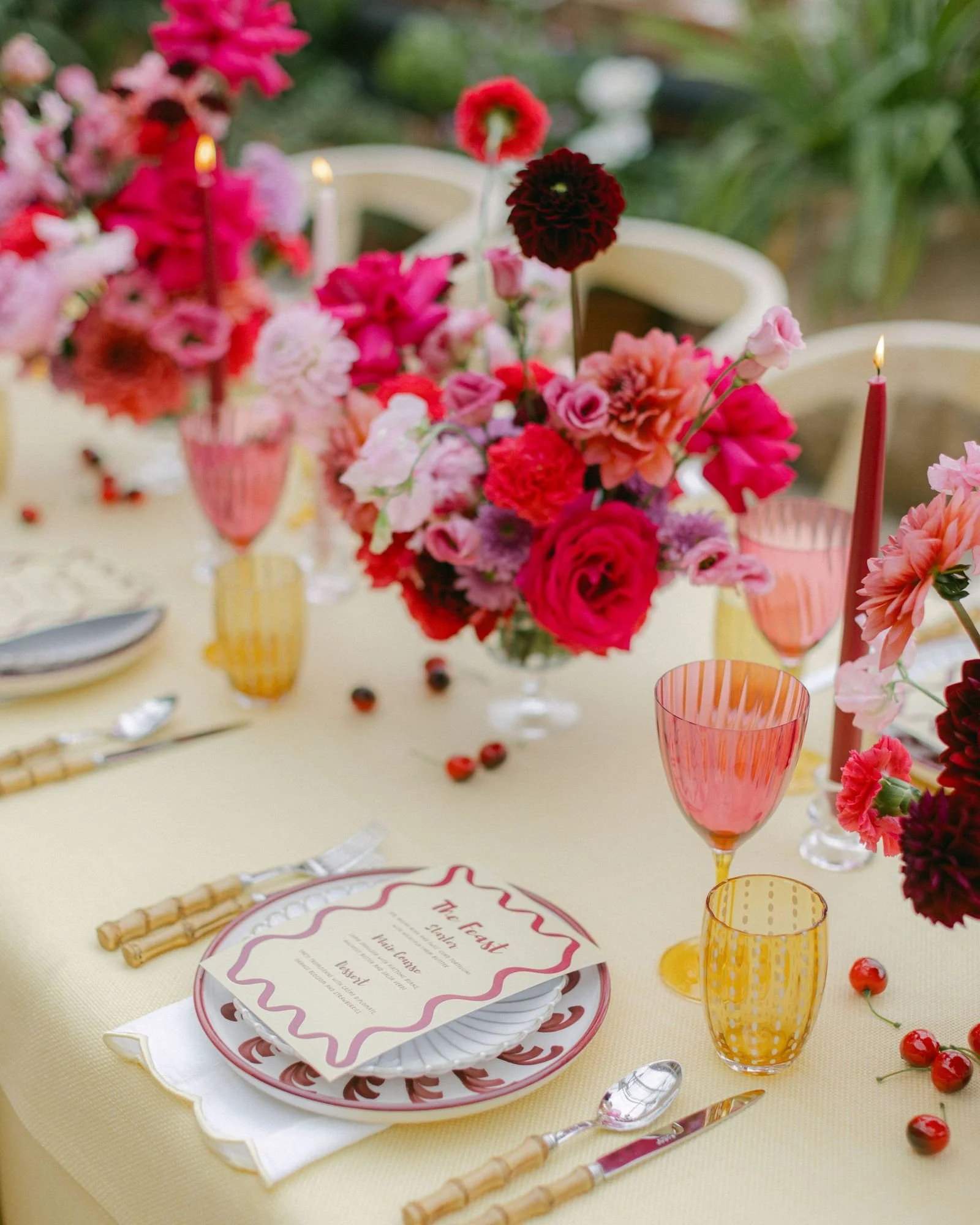 A table setting with colorful flower centerpieces, pink and yellow glasses, a white plate with a meal menu, silverware with bamboo handles, and scattered cherries on a cream tablecloth.