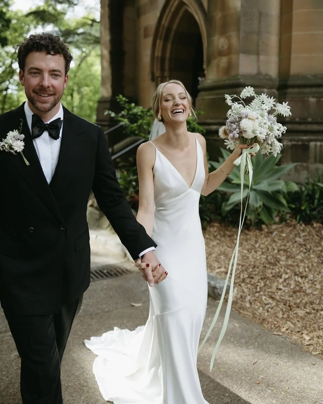 A newlywed couple holding hands, walking outdoors near a church, the bride smiling and holding a bouquet of white flowers, the groom dressed in a black tuxedo with a boutonniere, trees and stone building in the background.