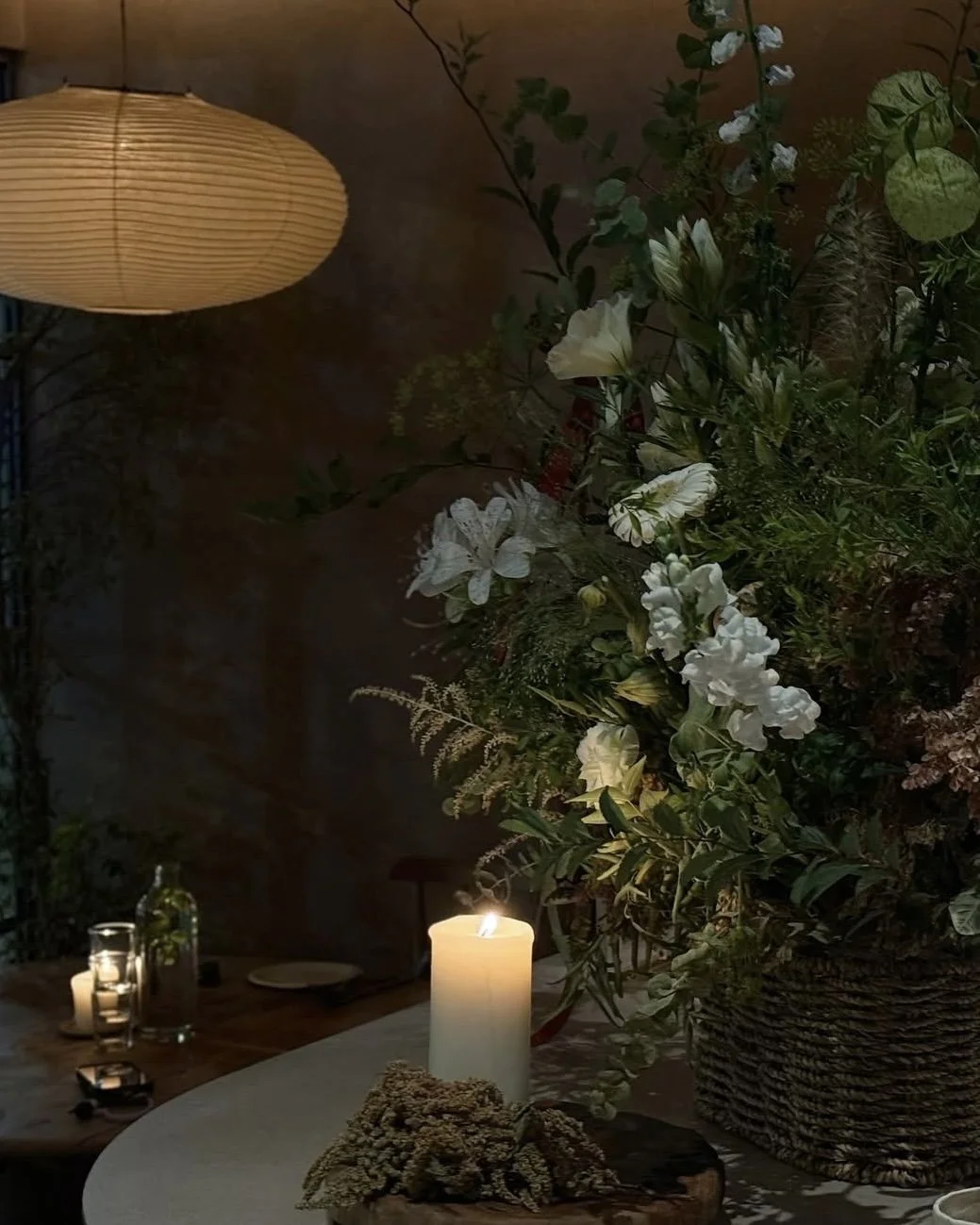 A table with a lit white candle, a large floral arrangement with white flowers and greenery, a wicker basket, and some smaller candles in glass holders. An overhead paper lantern provides warm lighting in a cozy indoor or outdoor setting.