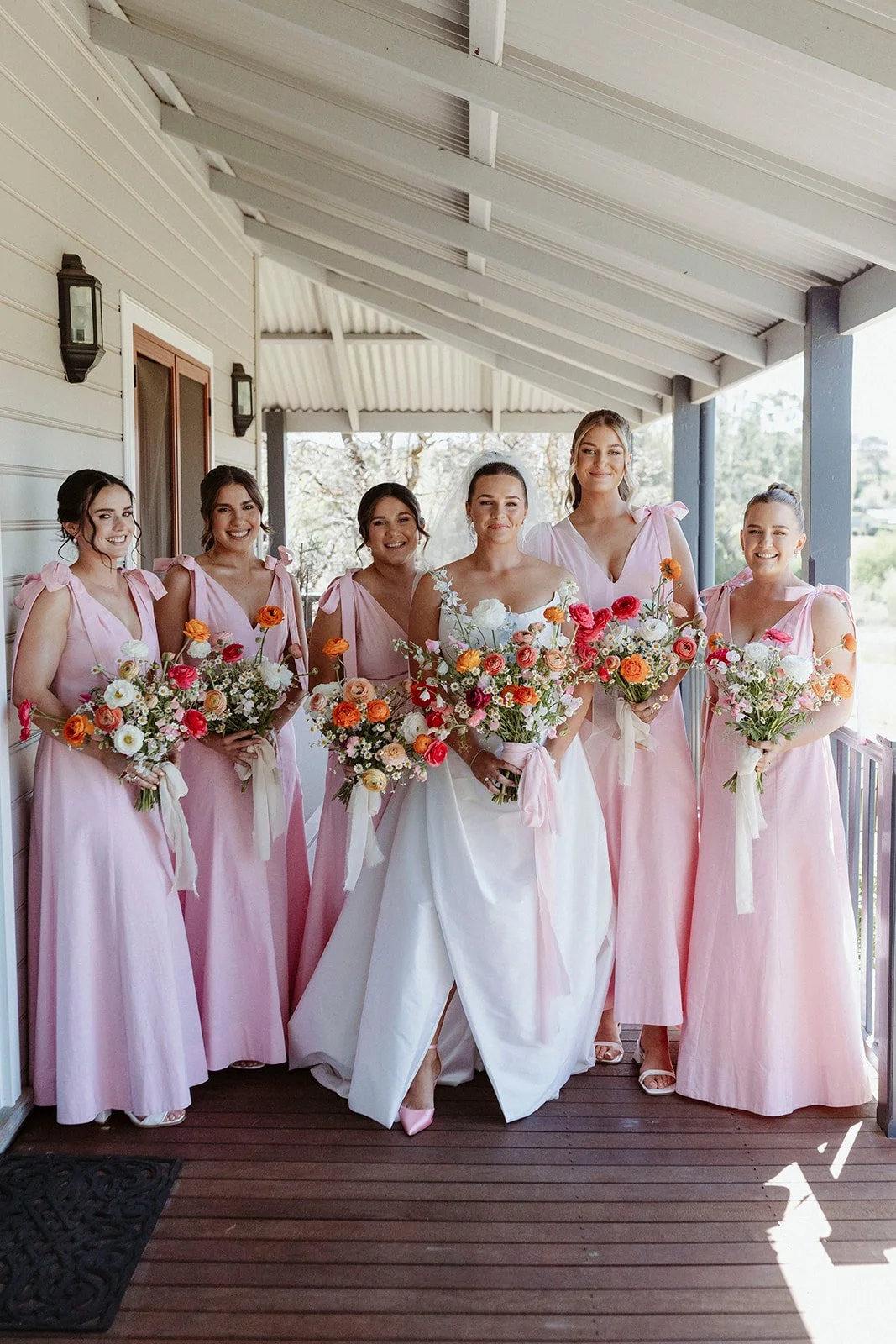 A bride and five bridesmaids standing on a porch, holding colorful bouquets of flowers, dressed in pink and white dresses, smiling for a photo.