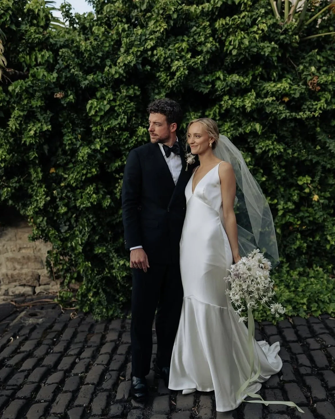 A bride and groom stand together outdoors in front of a dense green leafy wall, dressed in wedding attire. The bride wears a white satin wedding gown with a deep V neckline, holding a bouquet of white flowers, and has a long veil. The groom wears a b