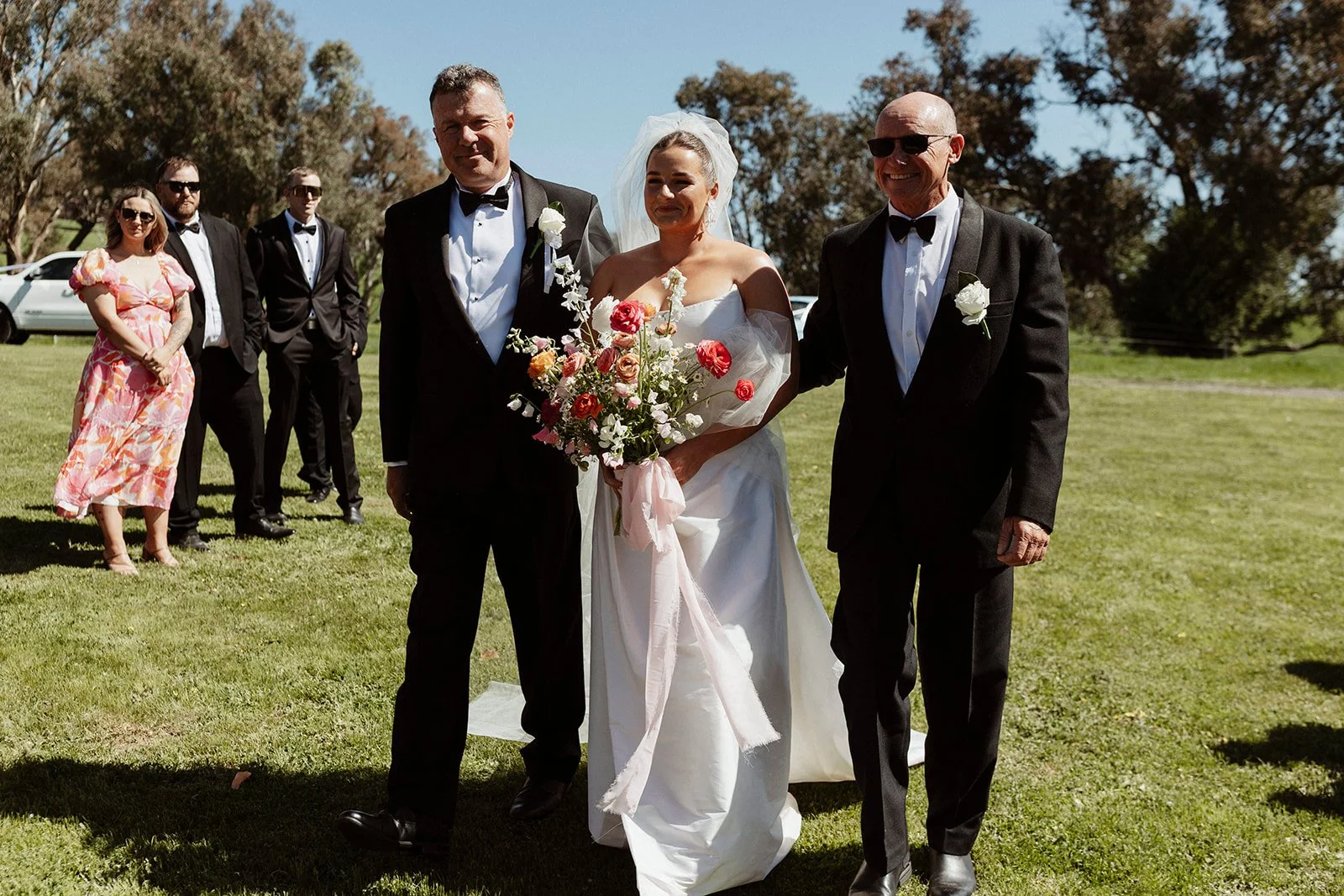 Wedding scene with bride in white gown holding flowers and two men in tuxedos, one on each side, walking on grass with guests in formal and casual attire in the background.