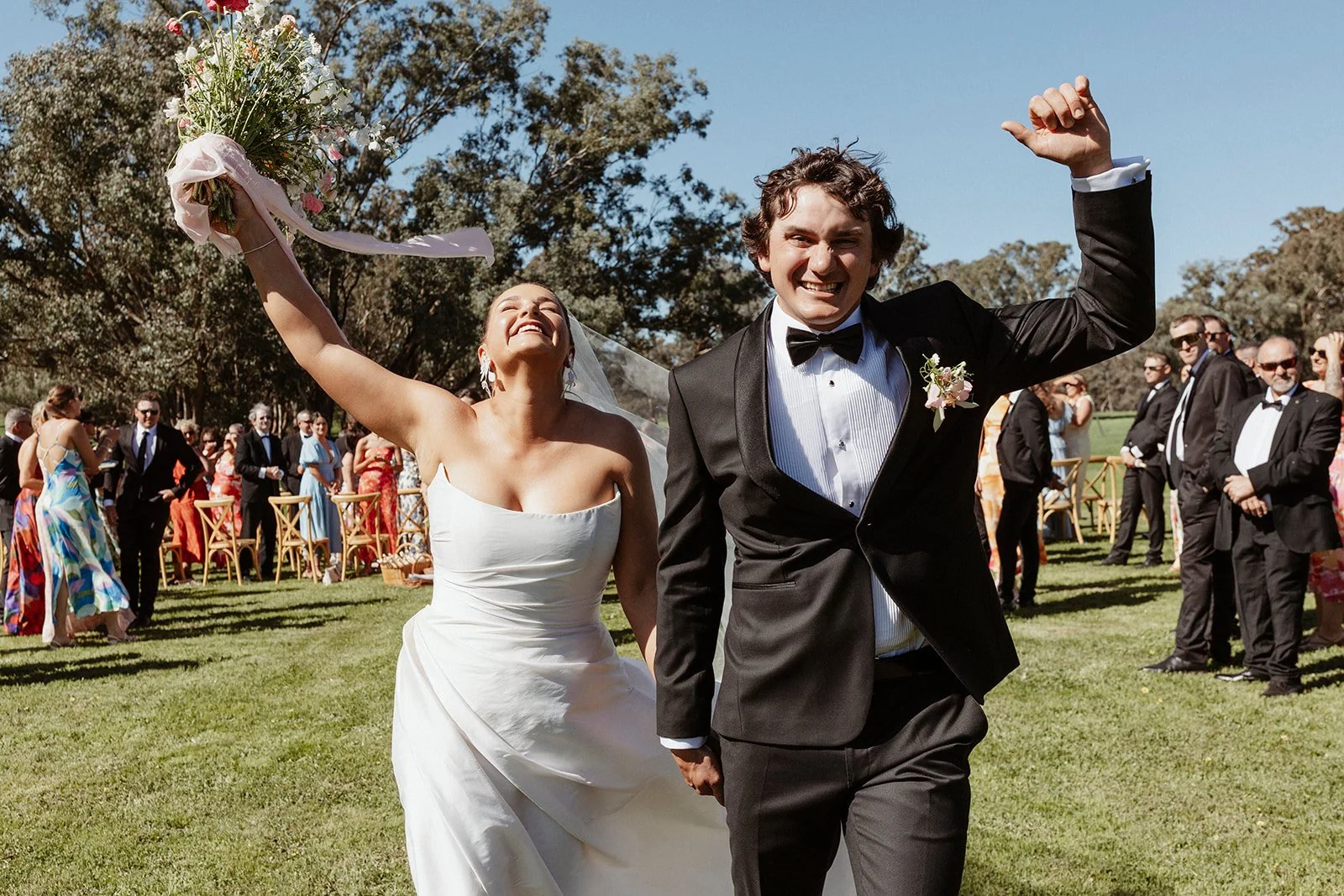 A newly married couple celebrating outdoors on a sunny day, with the bride holding a bouquet and the groom smiling and raising his fist in victory. Guests are in the background.