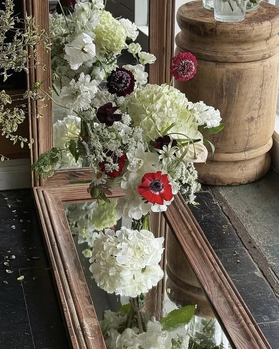 A floral arrangement of white, black, and red flowers resting on a mirror-topped wooden table, with a wooden container in the background.