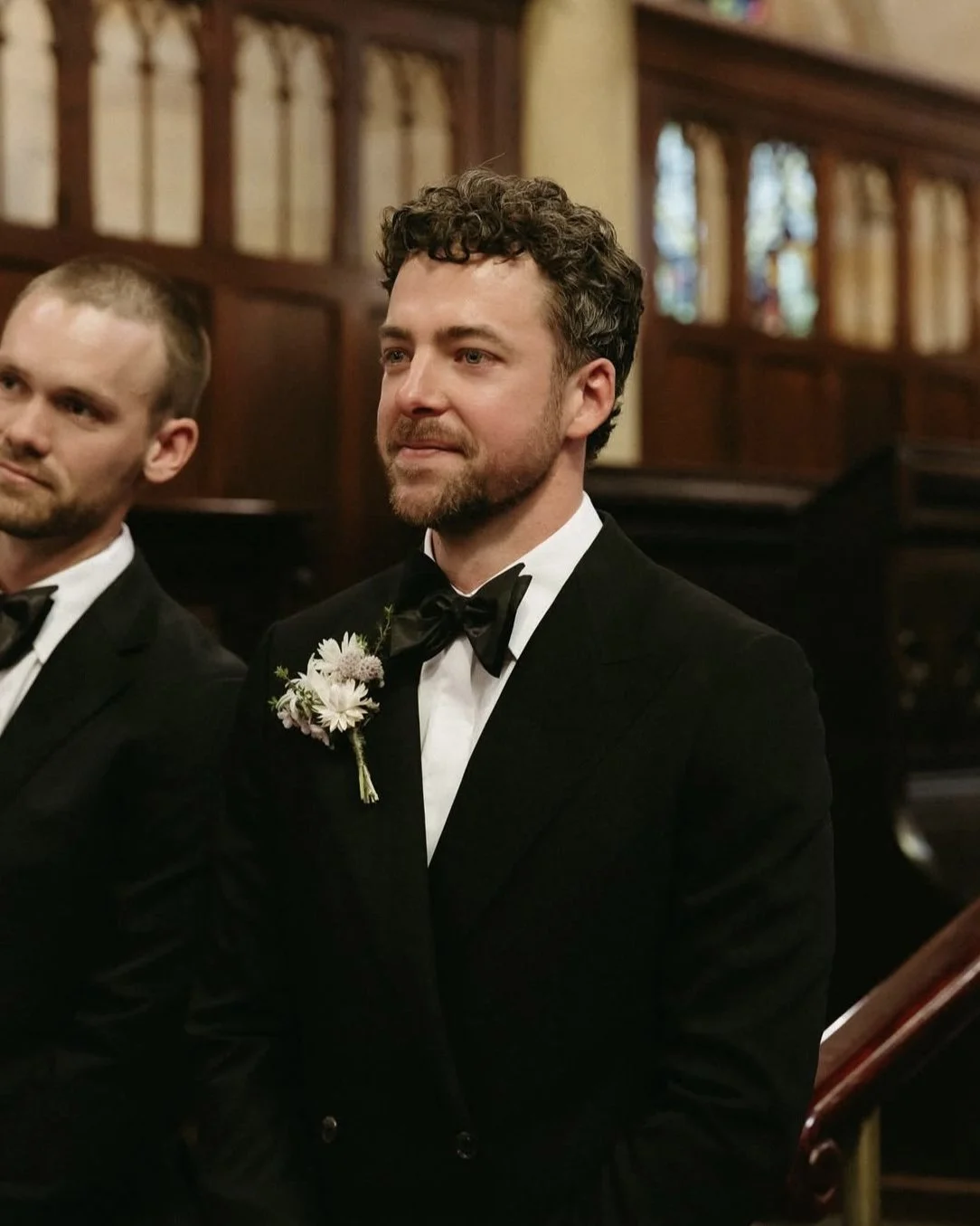 A man with curly hair and a beard wearing a tuxedo with a bow tie and a boutonniere standing indoors, possibly at a formal event or wedding.