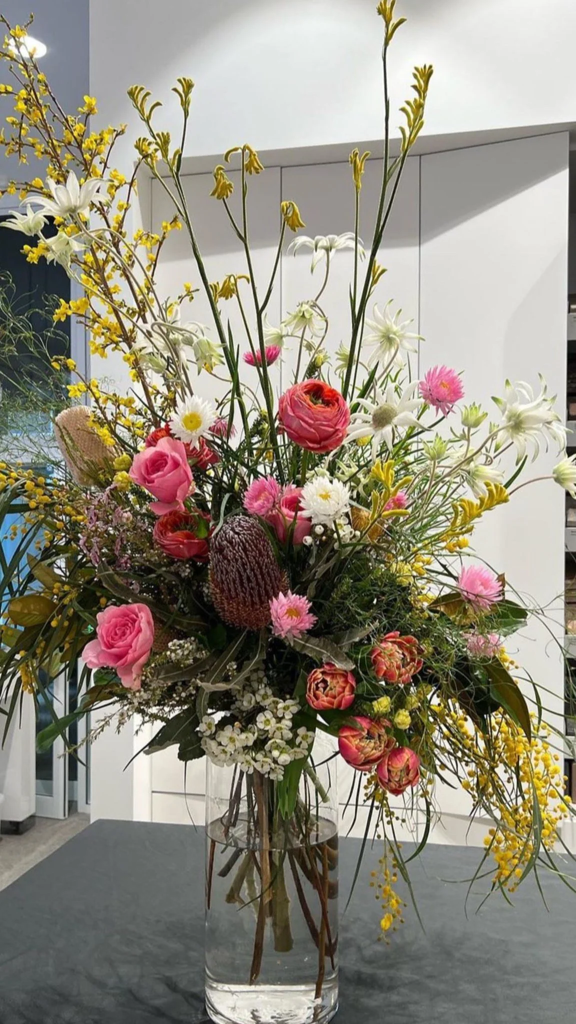 A colorful floral arrangement in a clear glass vase on a dark table, featuring pink roses, white lilies, yellow forsythia, and various other flowers and greenery.